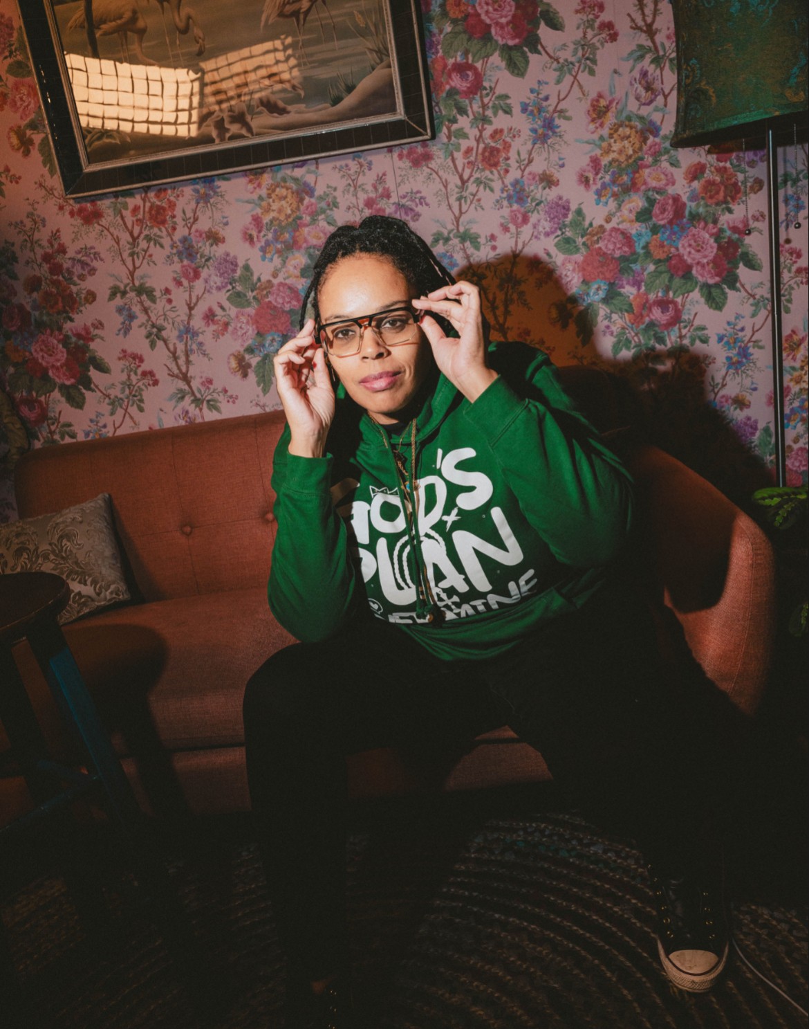 Person with glasses adjusting them, sitting on a sofa in a room with floral wallpaper, wearing a green hoodie.