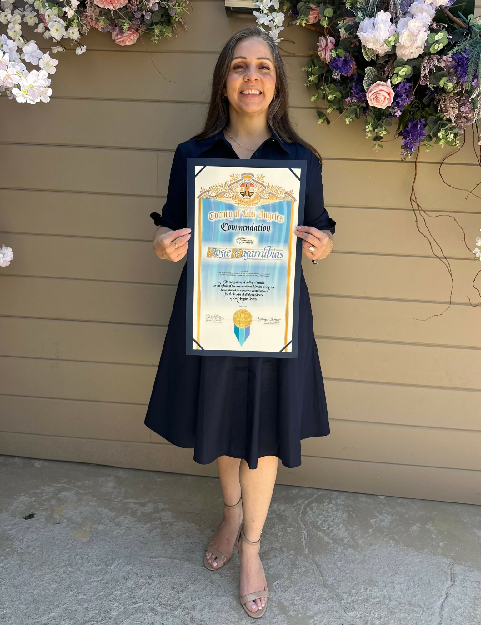 Young woman in a dark dress holding a framed certificate, standing in front of a floral backdrop, smiling.