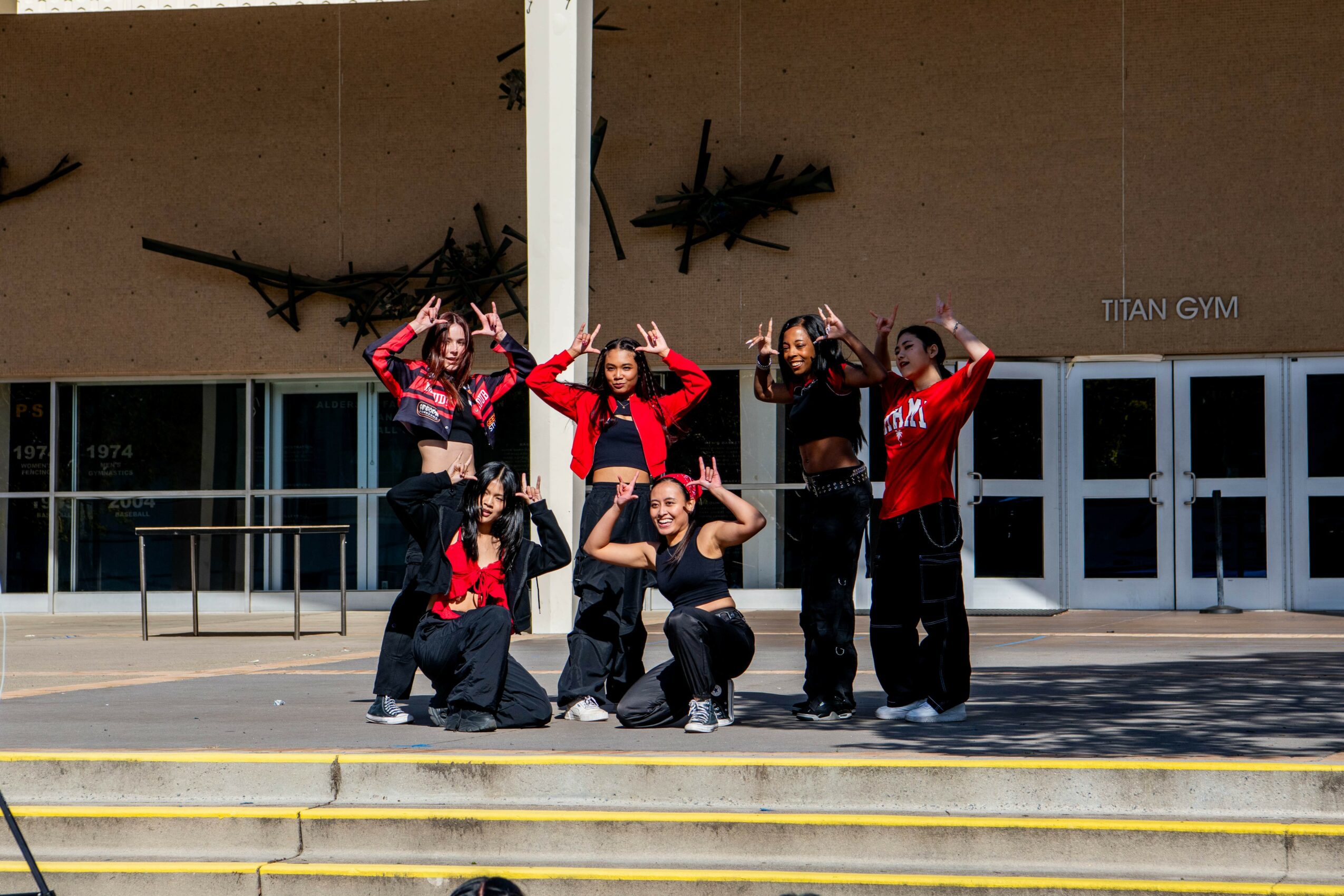 Group of six people posing outside a building with glass doors, some making peace signs, wearing black and red outfits.