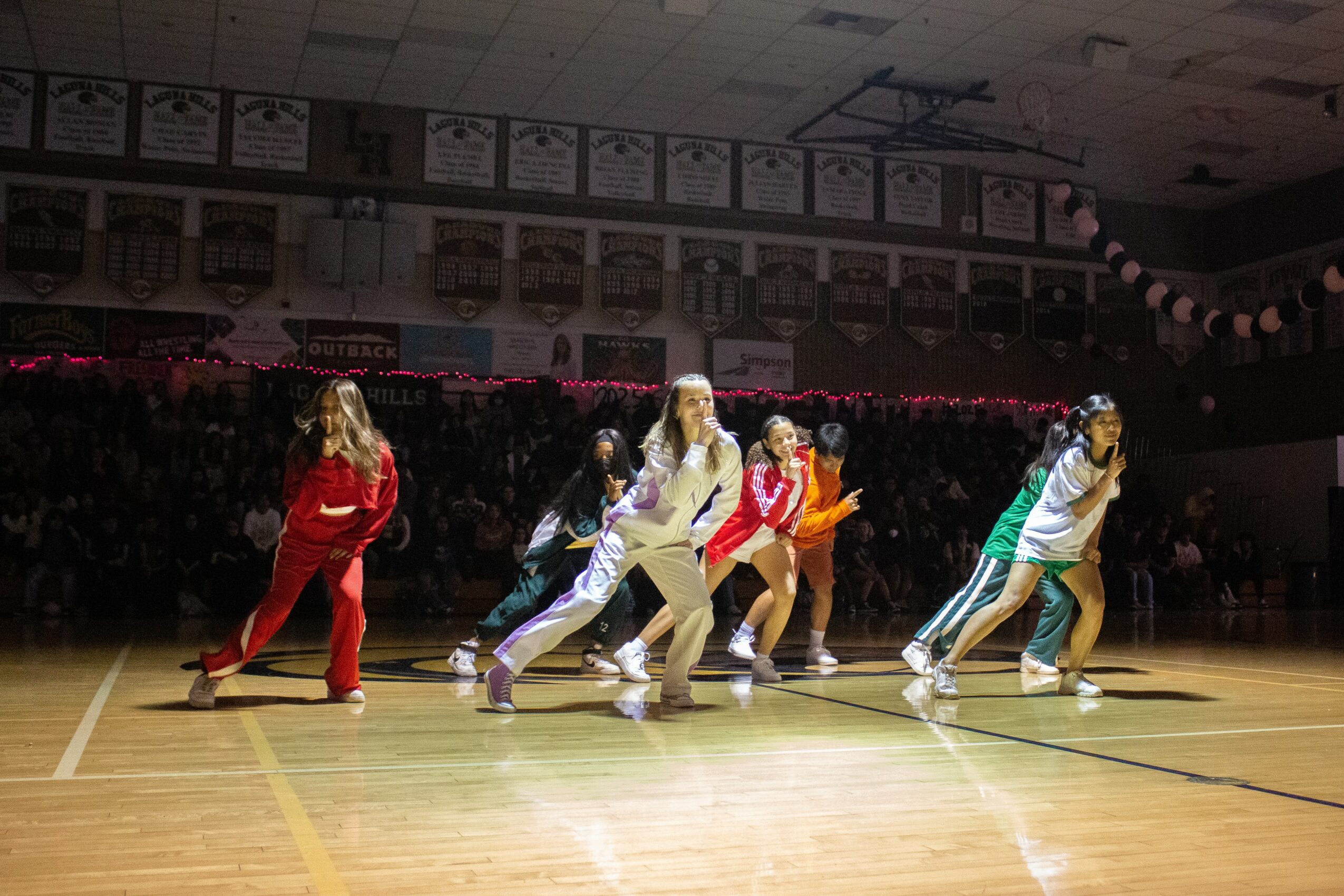 Group of people dancing on a gymnasium floor with an audience in the background, some wearing colorful clothing.