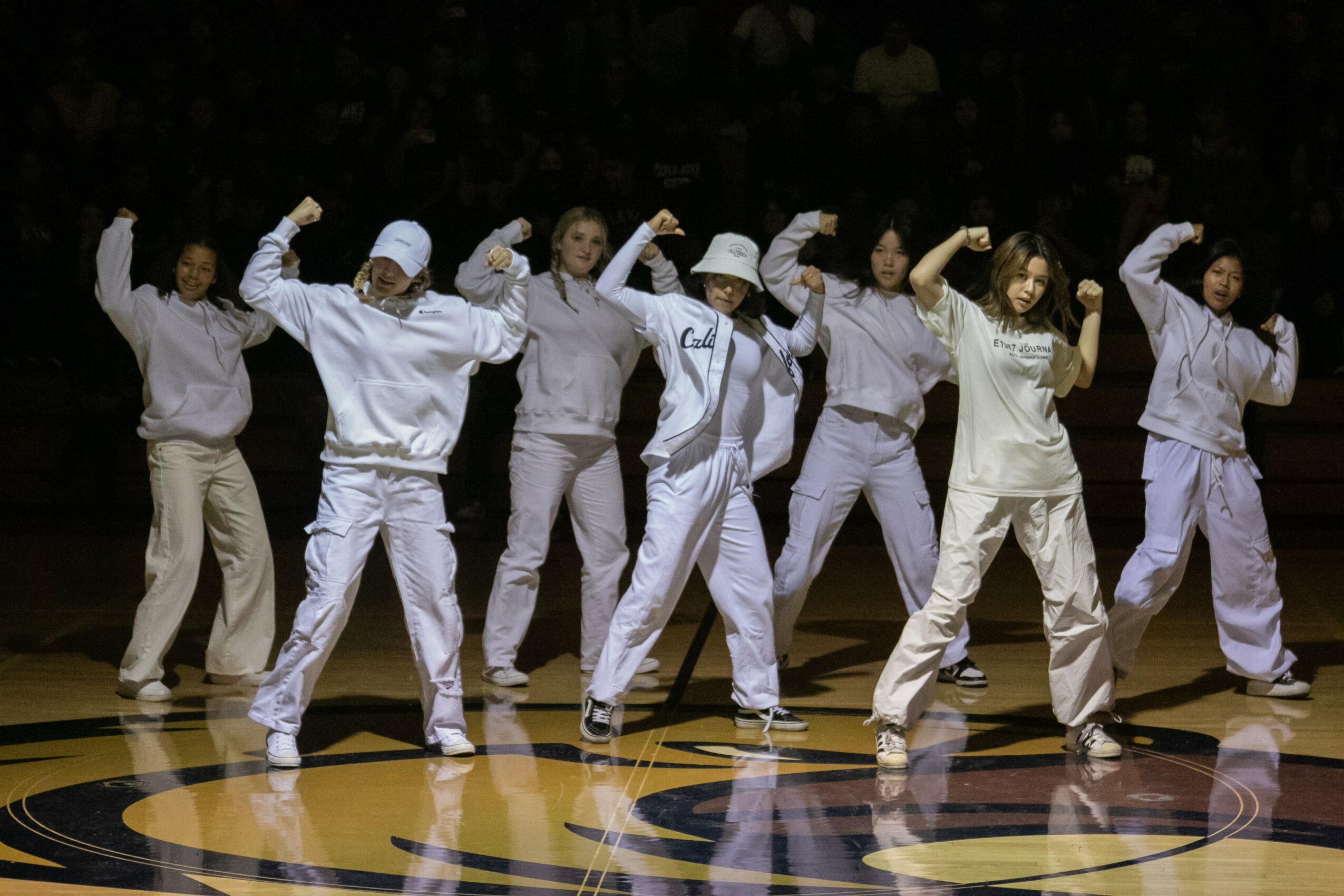 Group of eight people dancing on a stage, wearing white outfits, with arms raised and fists clenched, in a synchronized pose.