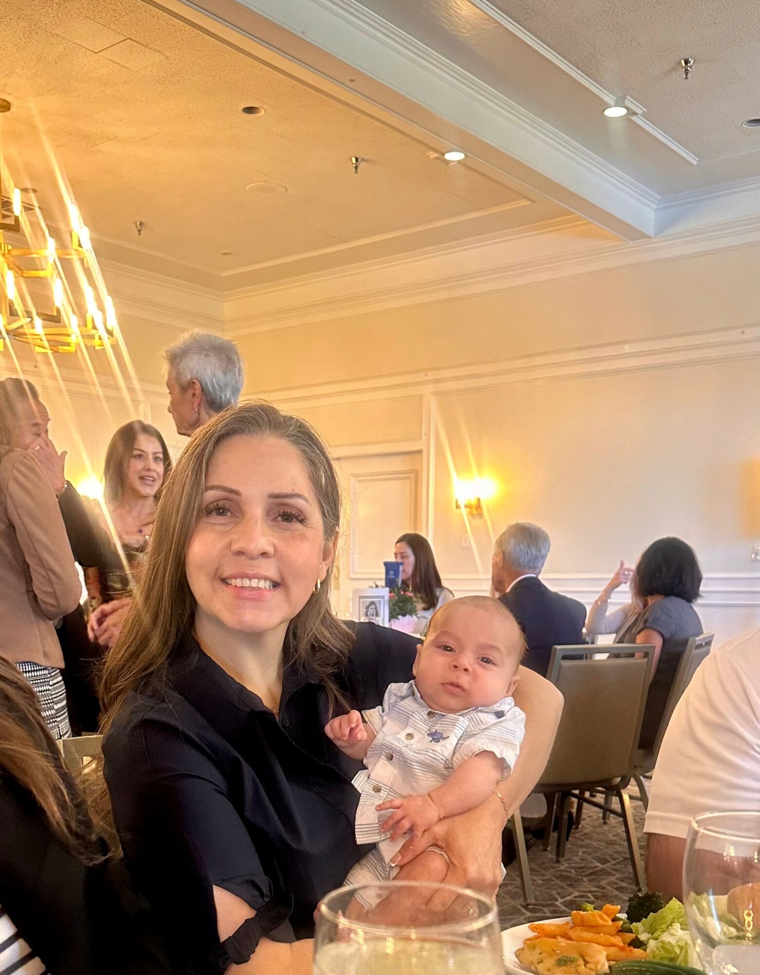 Woman holding a baby in a banquet hall with other people seated at tables, warm lighting, and elegant decor.