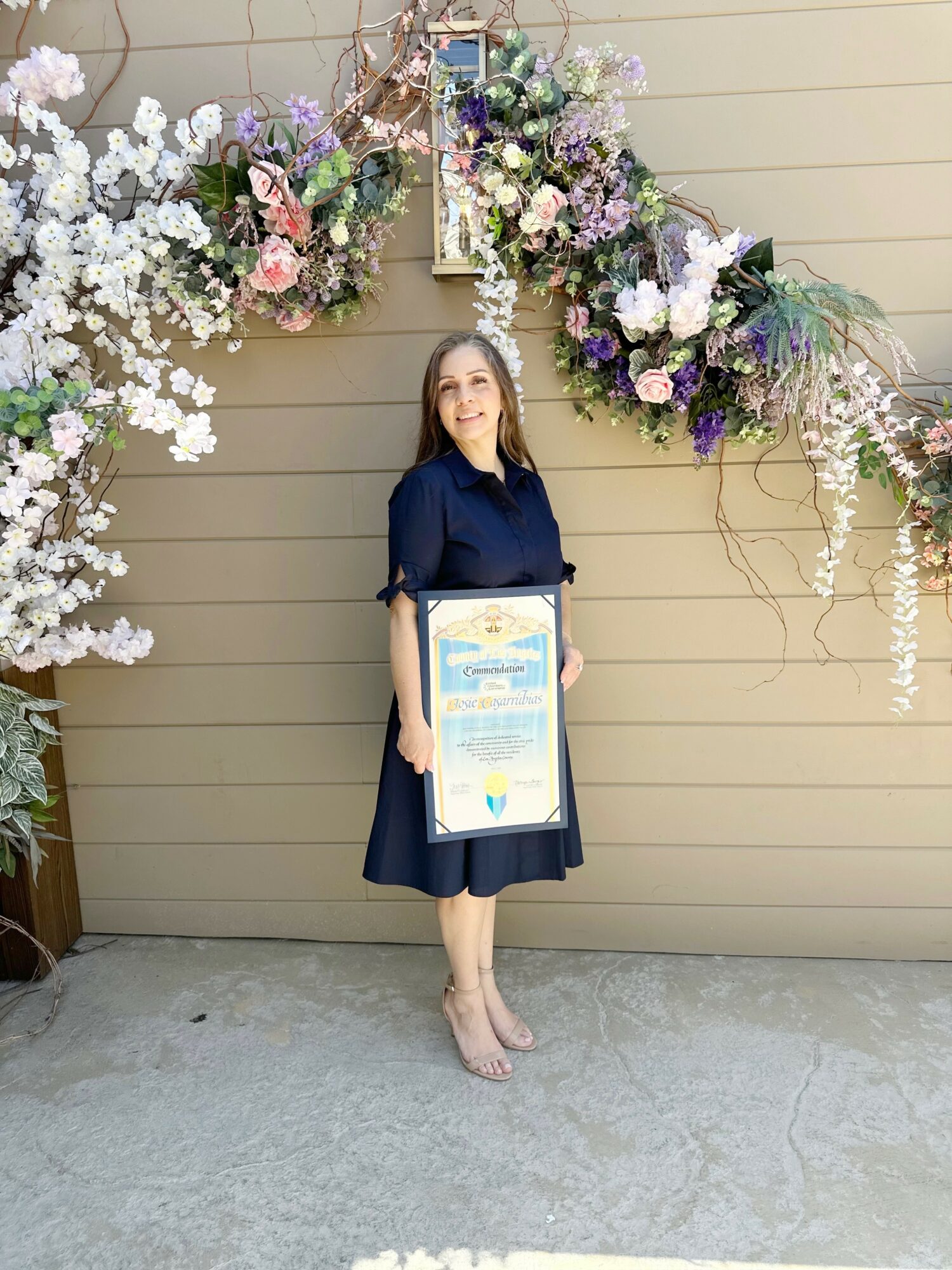 Woman in dark dress holding a framed certificate, standing in front of floral decorations on a beige wall.