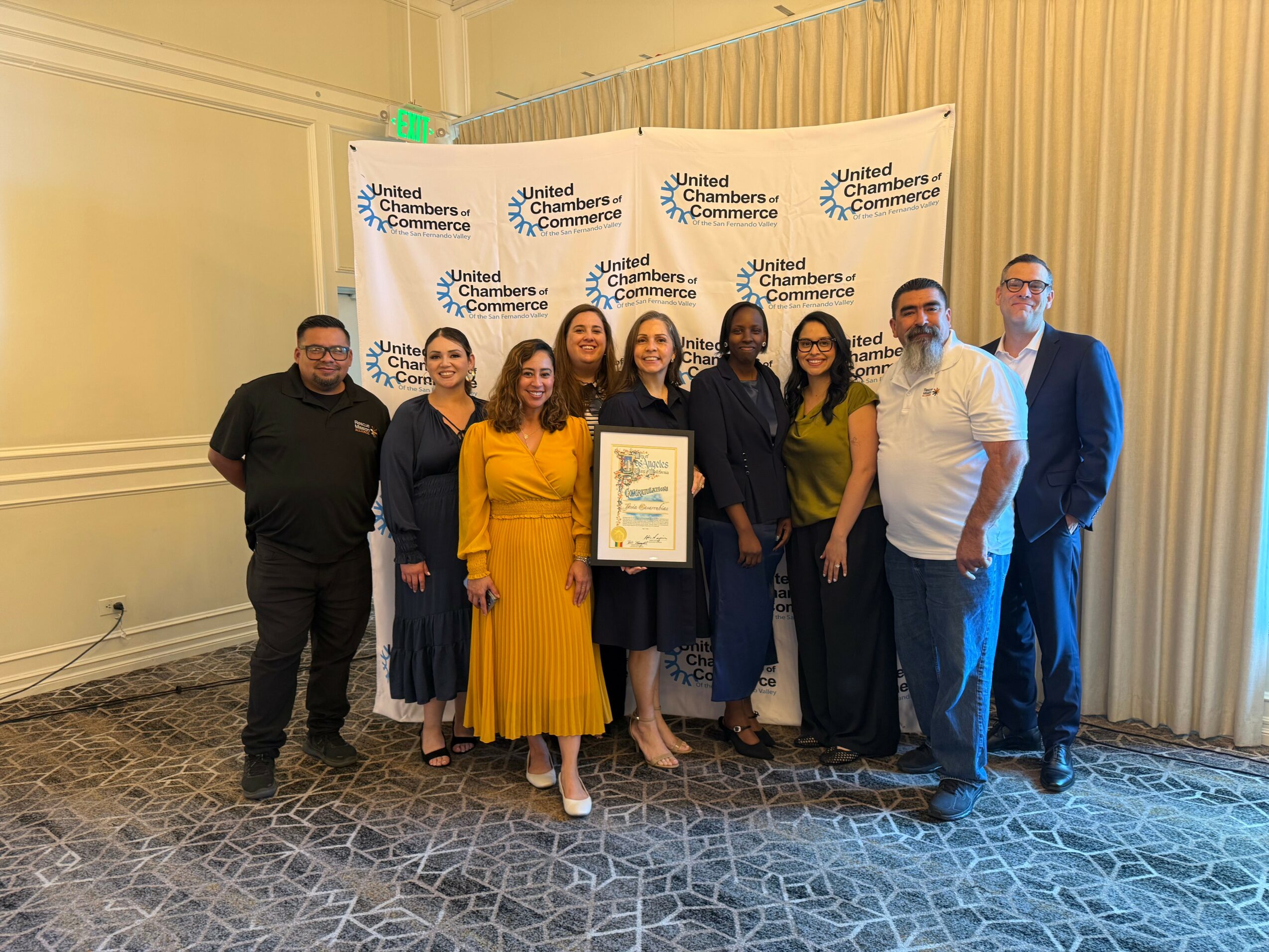 Group of nine people standing indoors, holding a framed certificate, in front of a backdrop with United Chamber of Commerce logos.