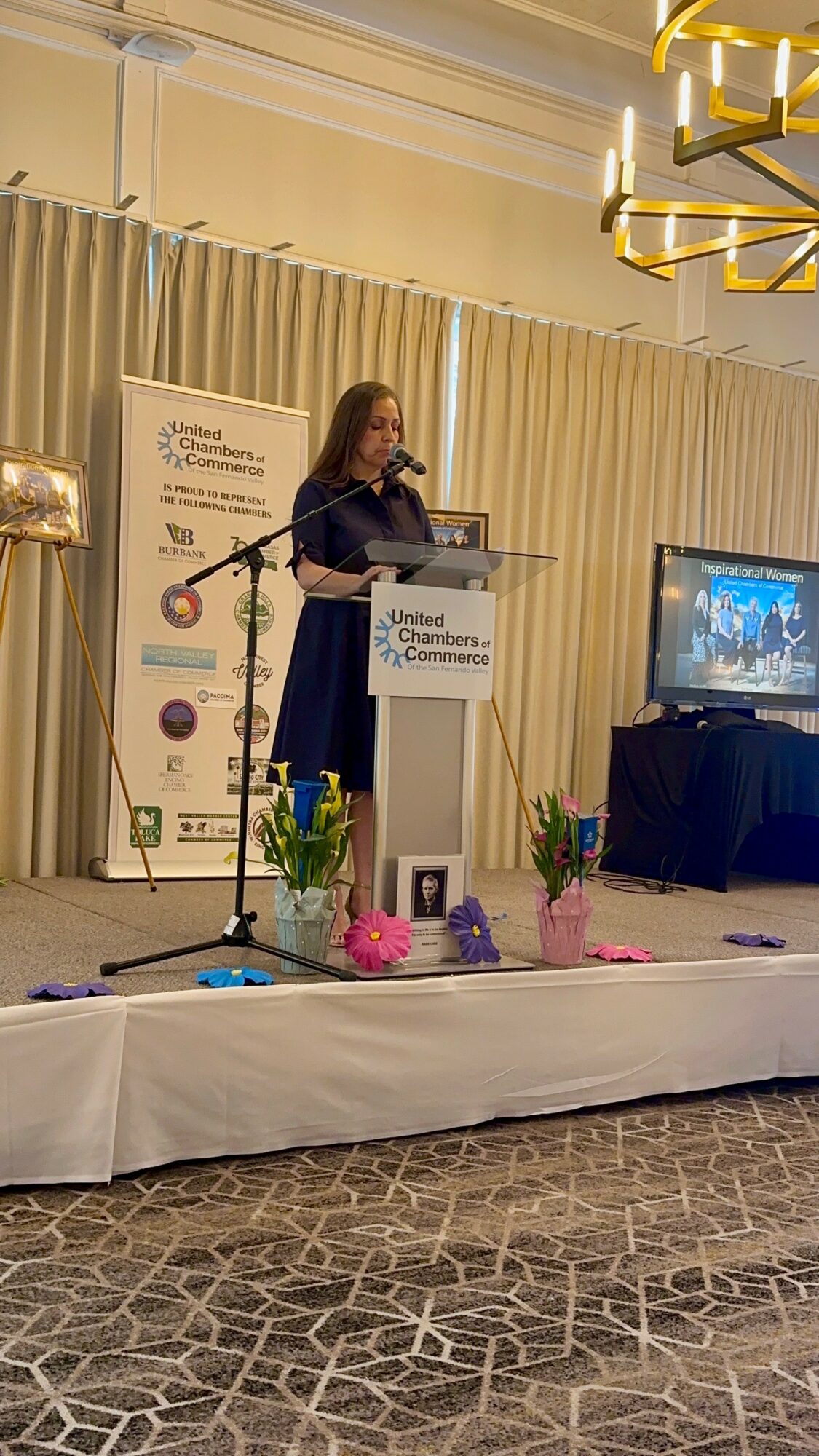 Woman speaking at a podium on stage with a screen displaying a presentation, decorated with flowers, in a conference room.