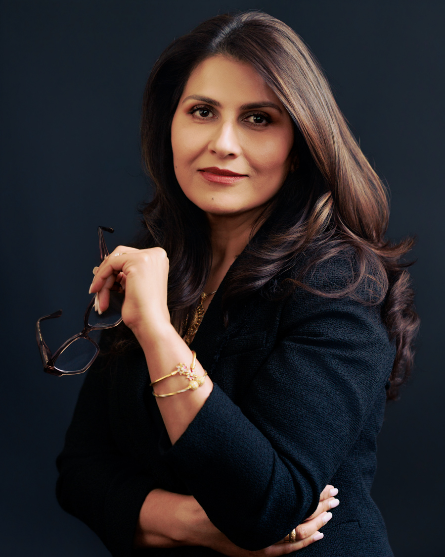 Woman with long dark hair, wearing a dark blazer, holding glasses, looking at camera, dark background.