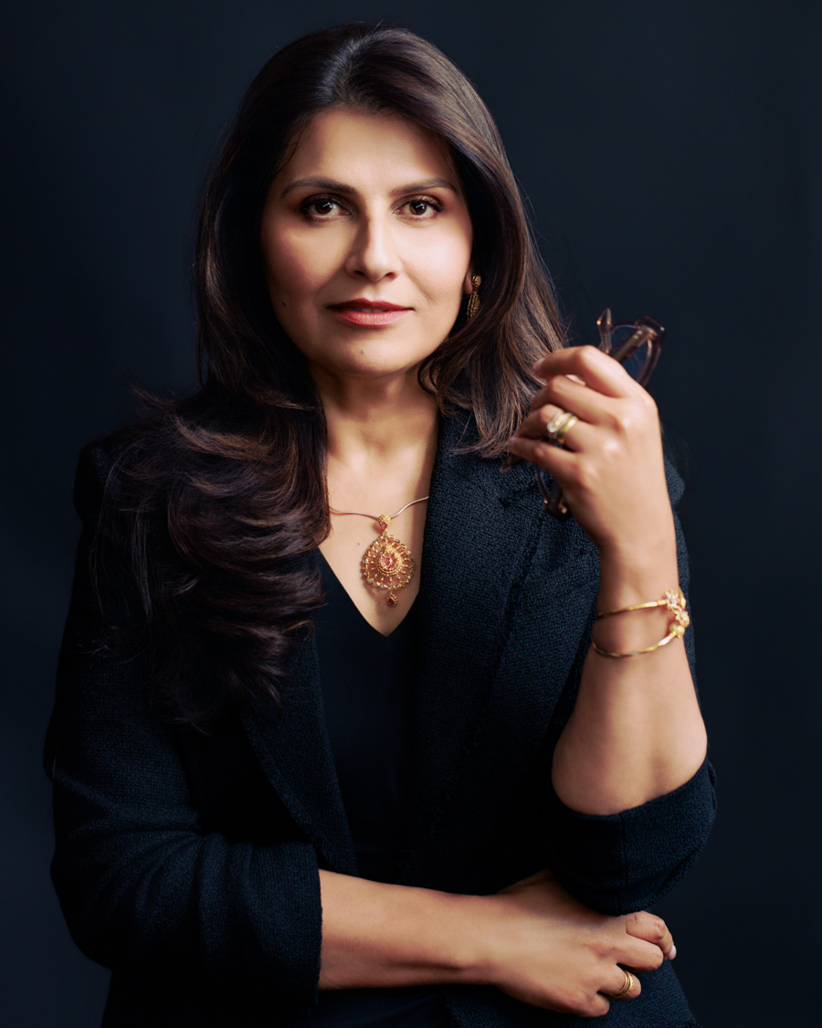 Woman with long dark hair wearing a black blazer and gold jewelry, looking at the camera against a dark background.