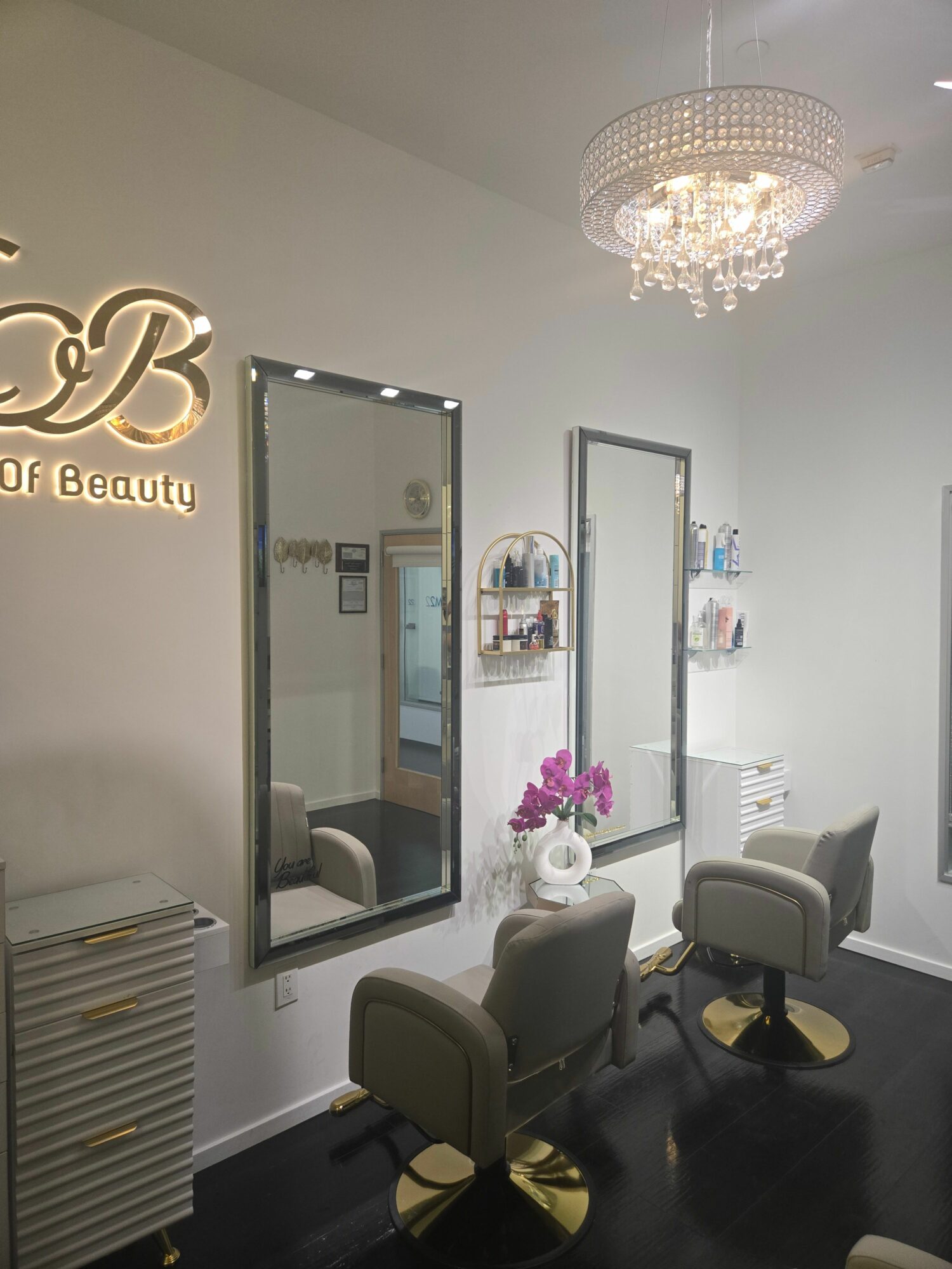 Interior of a beauty salon with mirrors, chairs, a chandelier, and a flower arrangement on a table.