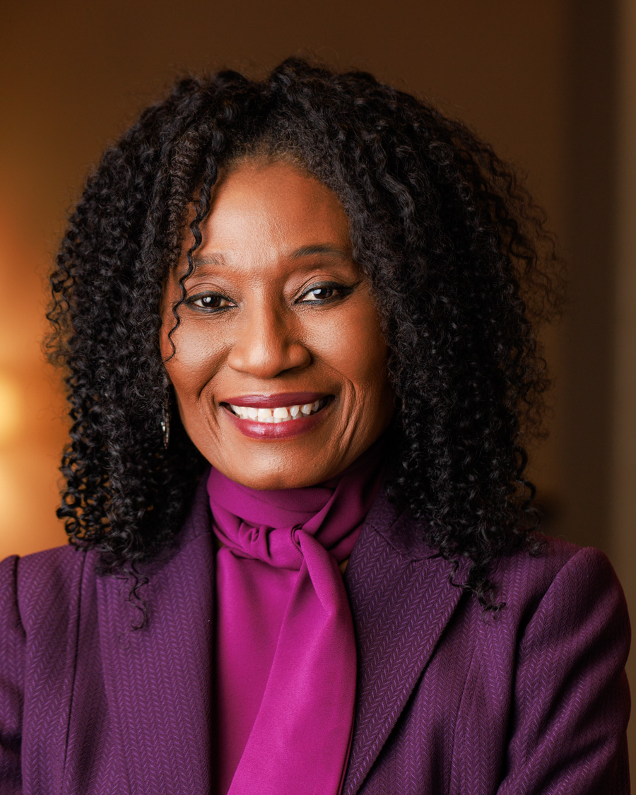 Portrait of a woman with curly hair smiling, wearing a purple blazer and matching scarf, against a warm background.