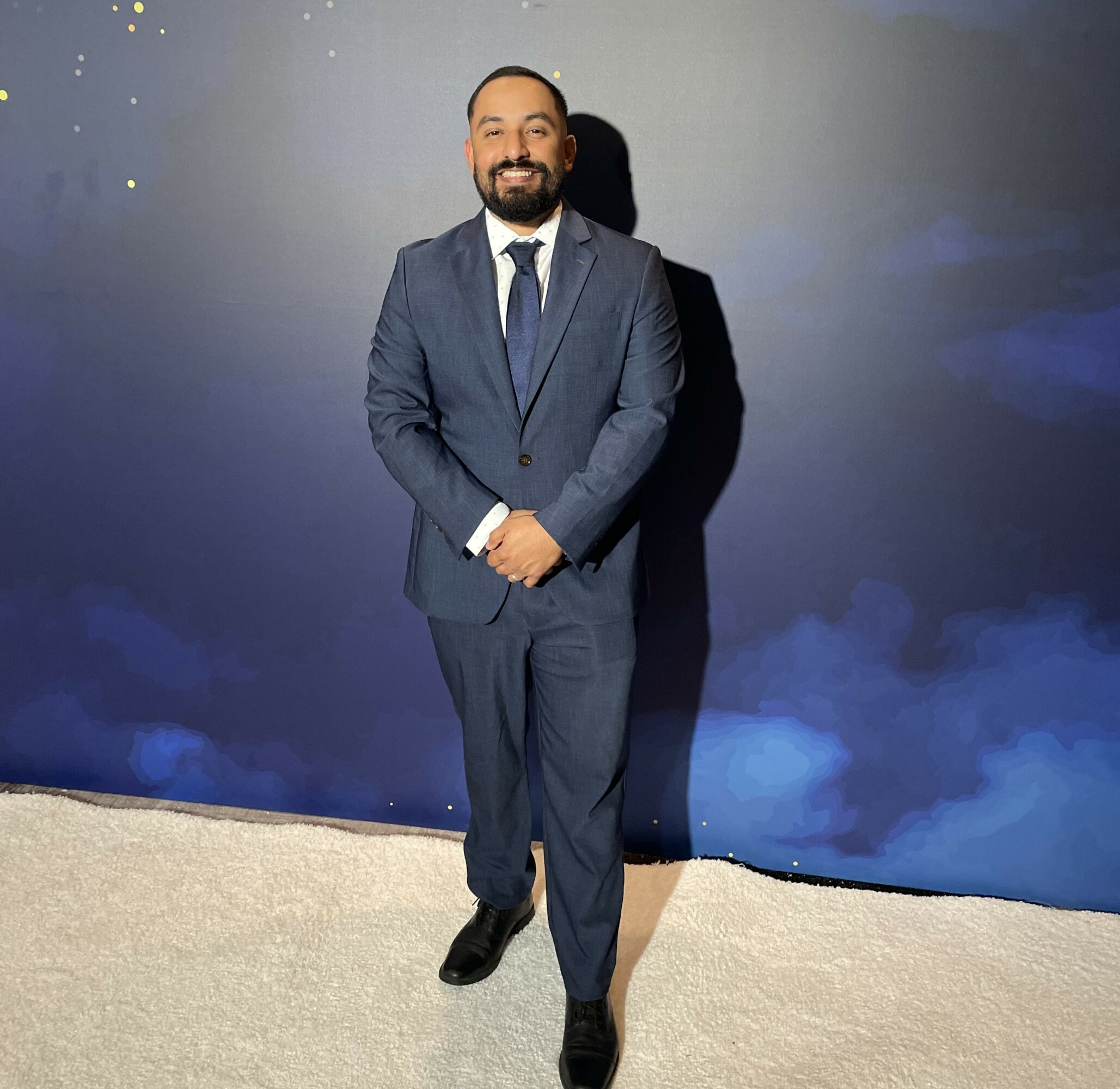 Man in a suit standing on a beige carpet, smiling, with a dark blue background. Word count: 20