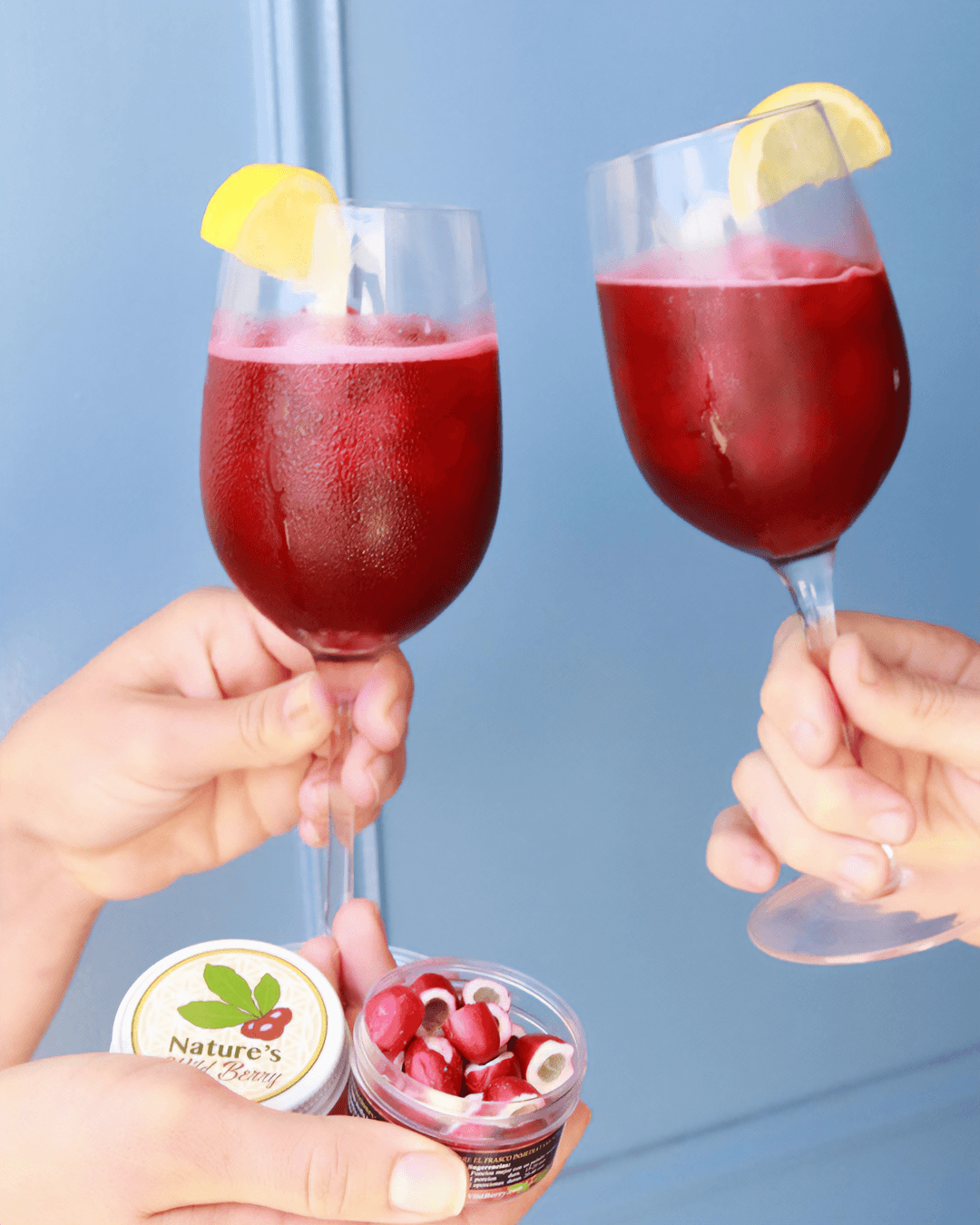 Two hands hold glasses of red beverage with lemon slices, one hand also holds a small bowl of berries, against a blue background.
