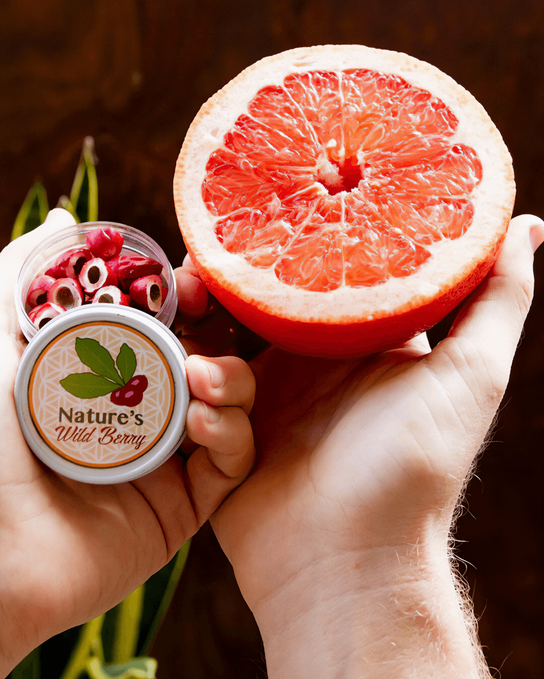 Person holding a halved grapefruit and a small container of berry-flavored lip balm, with pink flowers in the background.
