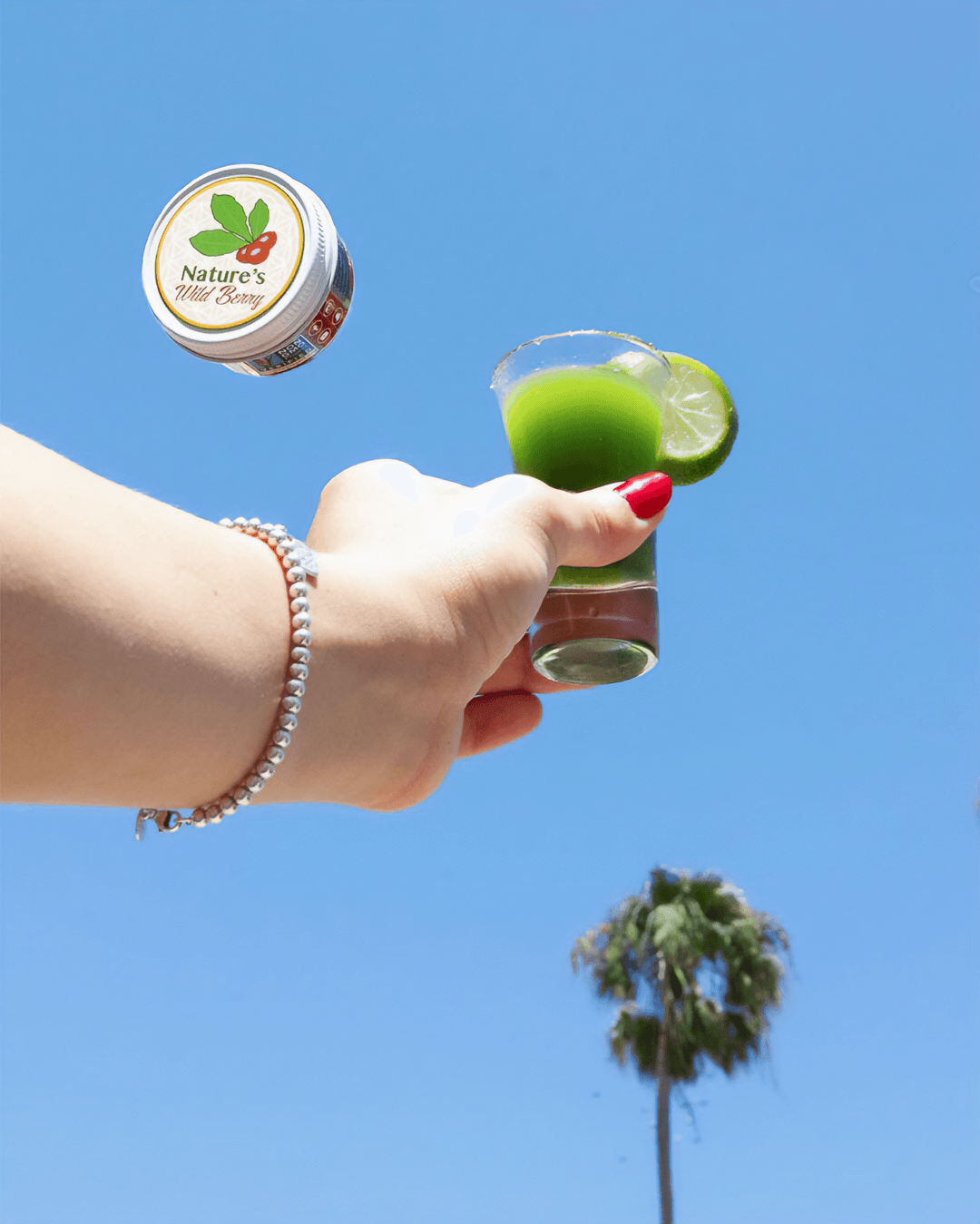 Hand holding a shot glass with green liquid and lime, against a blue sky with a palm tree, floating container nearby.