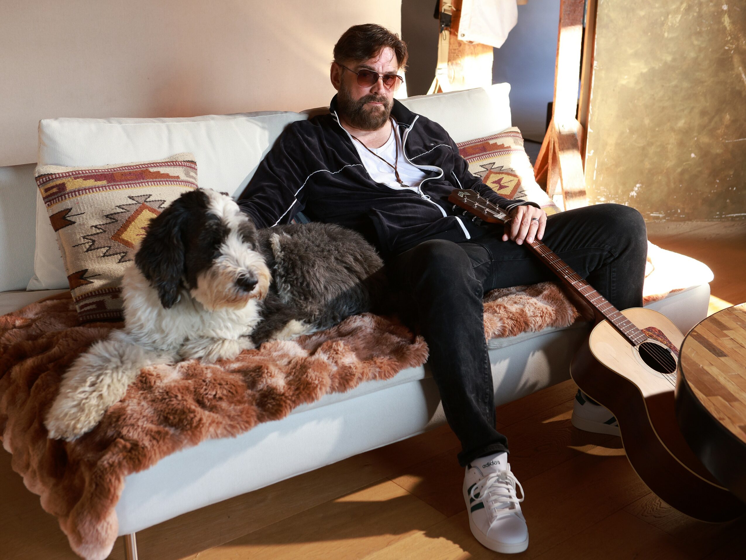 Man with sunglasses and beard sitting on a sofa with a large dog, guitar nearby, in a cozy room.