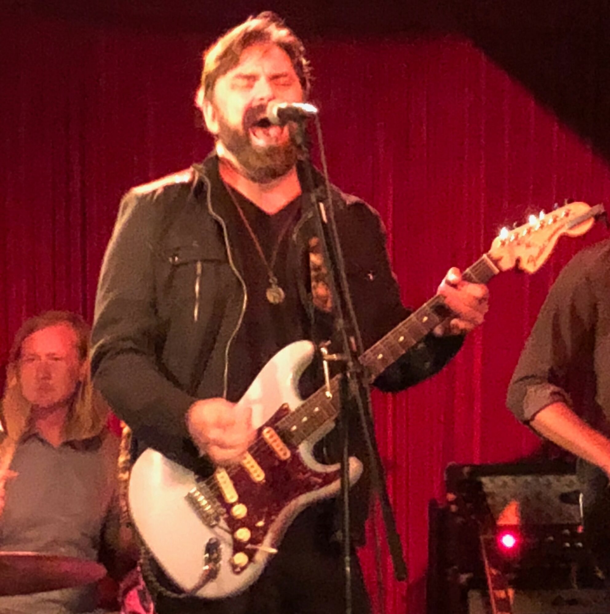 Man singing and playing electric guitar on stage with red curtain background.
