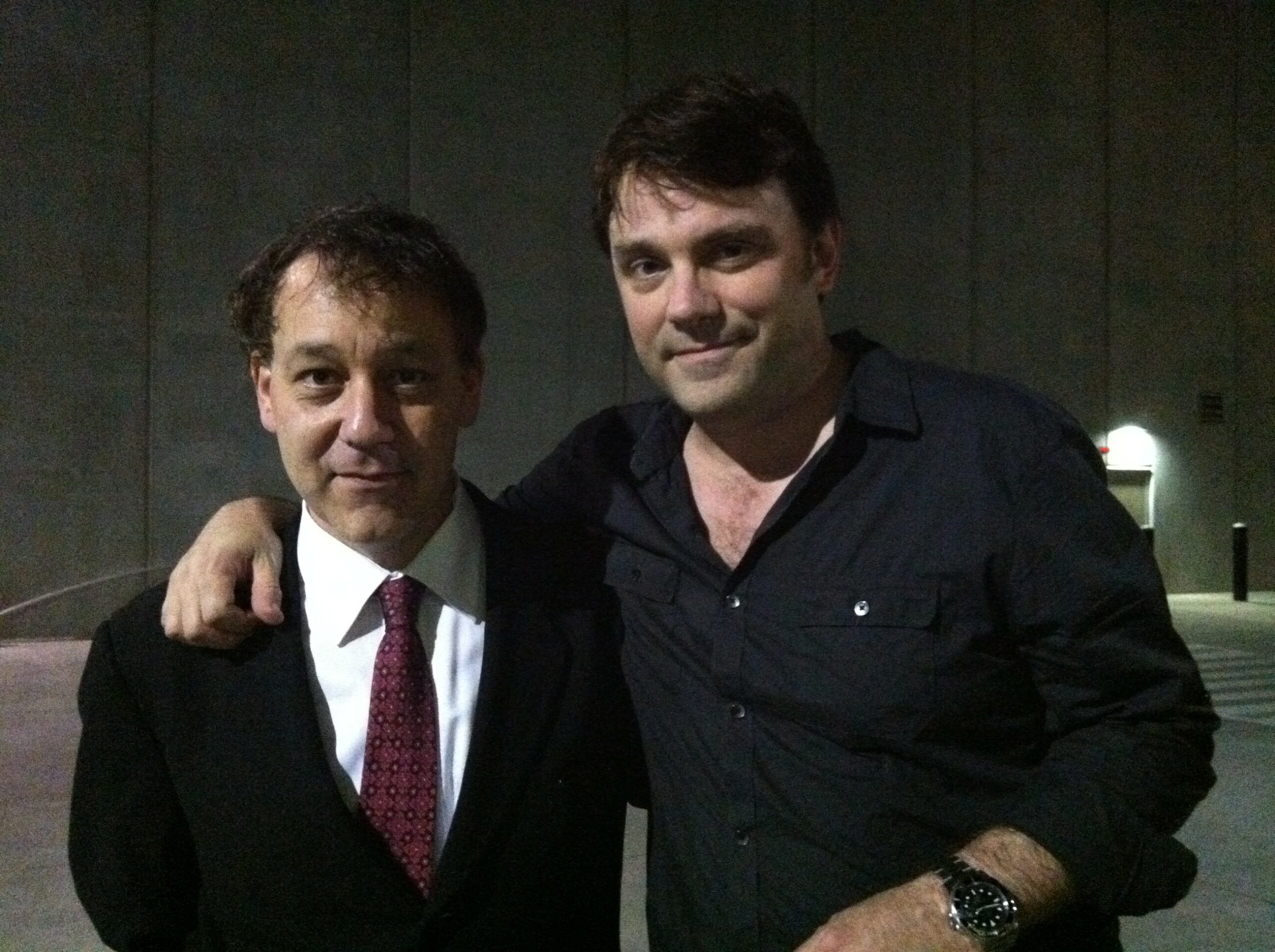 Two men stand close together outdoors at night, one with arm around the other, both smiling.