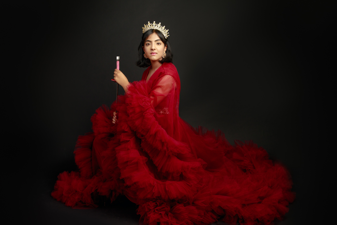 Woman in red gown with a crown, holding a wand, sitting against a dark background.