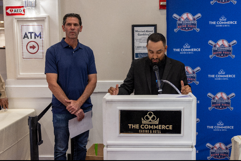 Two men stand in front of a podium with a sign reading 'The Commerce.' One man is speaking at the podium, the other stands nearby holding papers.