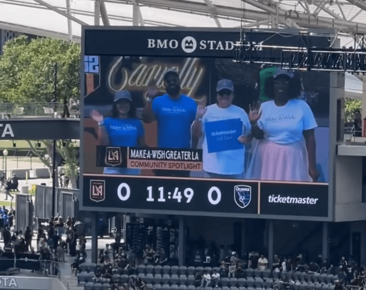 Large digital scoreboard showing a group of people holding signs, with a stadium and trees in the background.