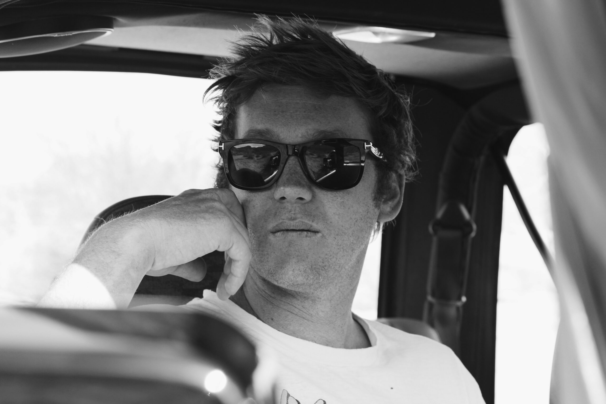 Man wearing sunglasses sitting in vehicle, resting his chin on his hand, looking forward, black and white photo.