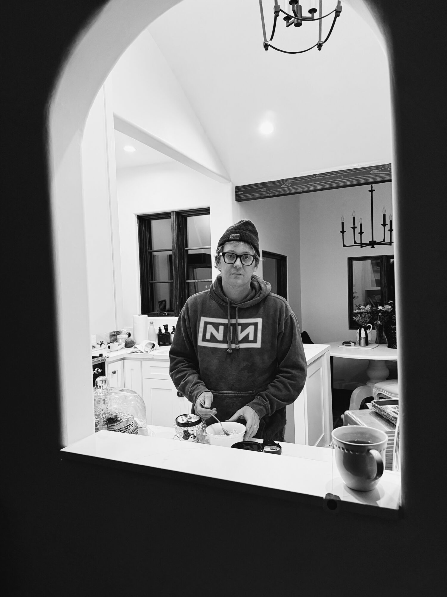 Young person with glasses and hoodie standing in kitchen, looking at camera, reflected in mirror, with kitchen items visible.