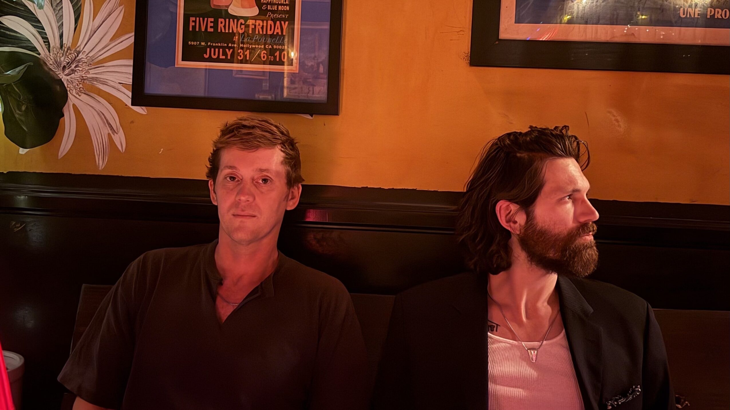 Two men sitting indoors against a yellow wall with framed posters, one with short hair and the other with longer hair and a beard.