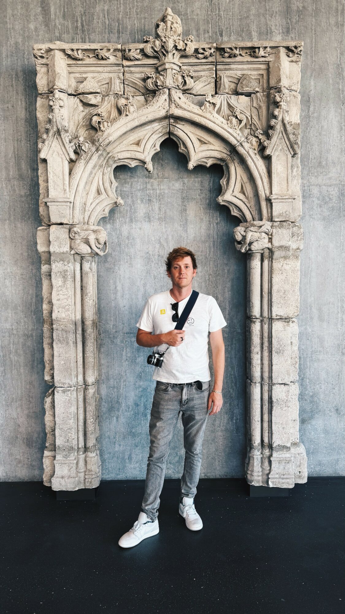 Young man standing in front of an ornate stone archway, wearing a white t-shirt, jeans, and white sneakers, holding a camera strap.