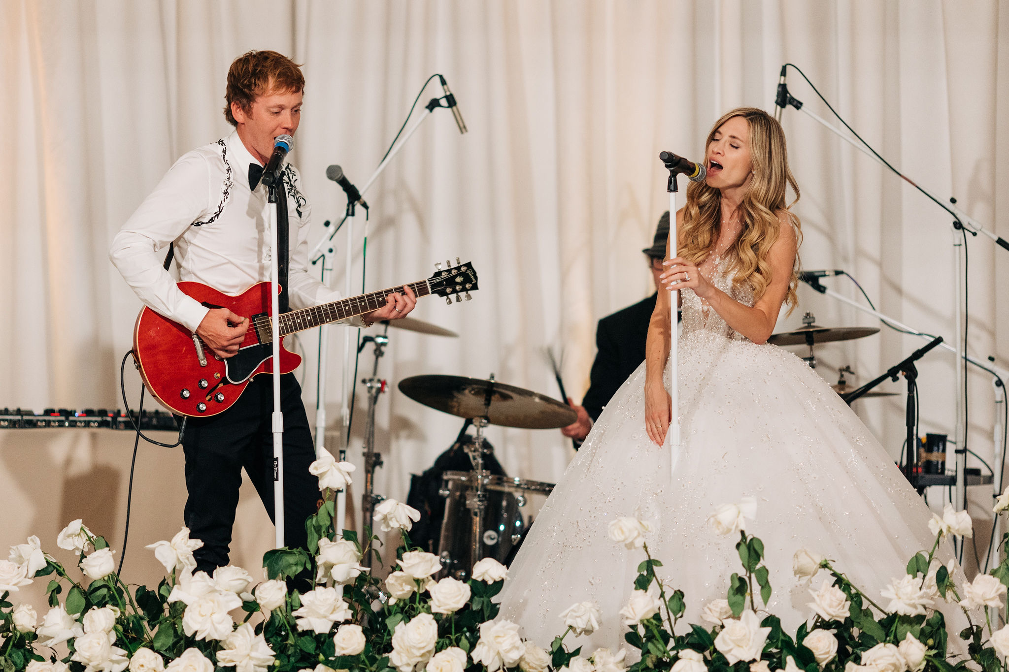 A young man and woman perform on stage with musical instruments, surrounded by white flowers and curtains.