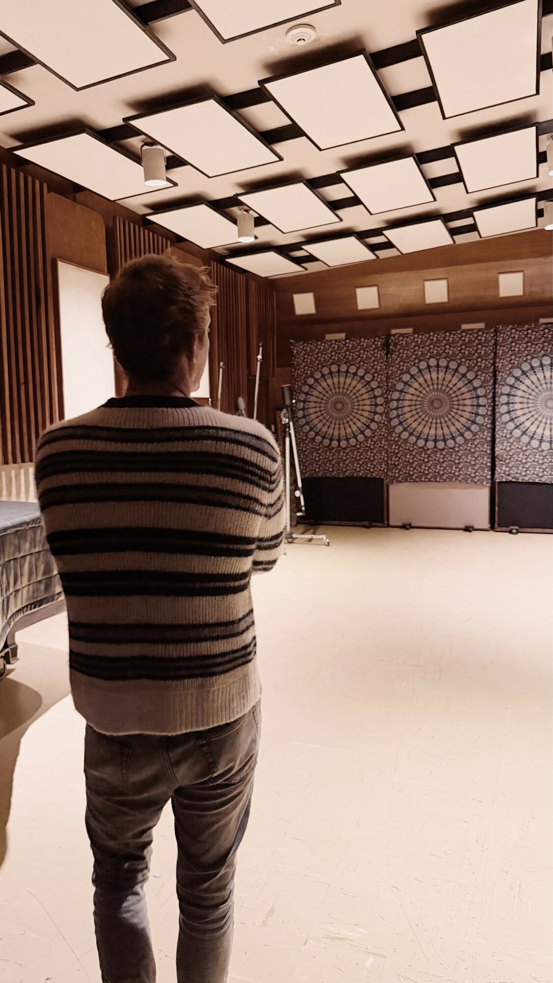 Person with short hair wearing a striped sweater and pants, standing in a room with patterned walls and ceiling lights.