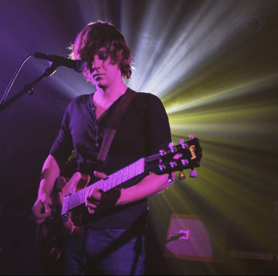 Person playing guitar on stage with colorful lighting behind them.