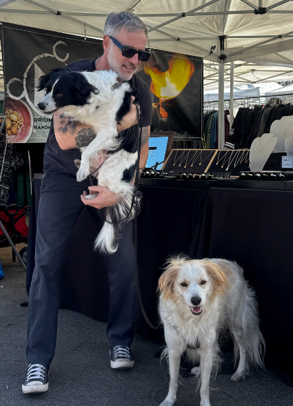 Person holding a black and white dog, standing outdoors near a booth with a banner and clothing racks, another dog on a leash nearby.