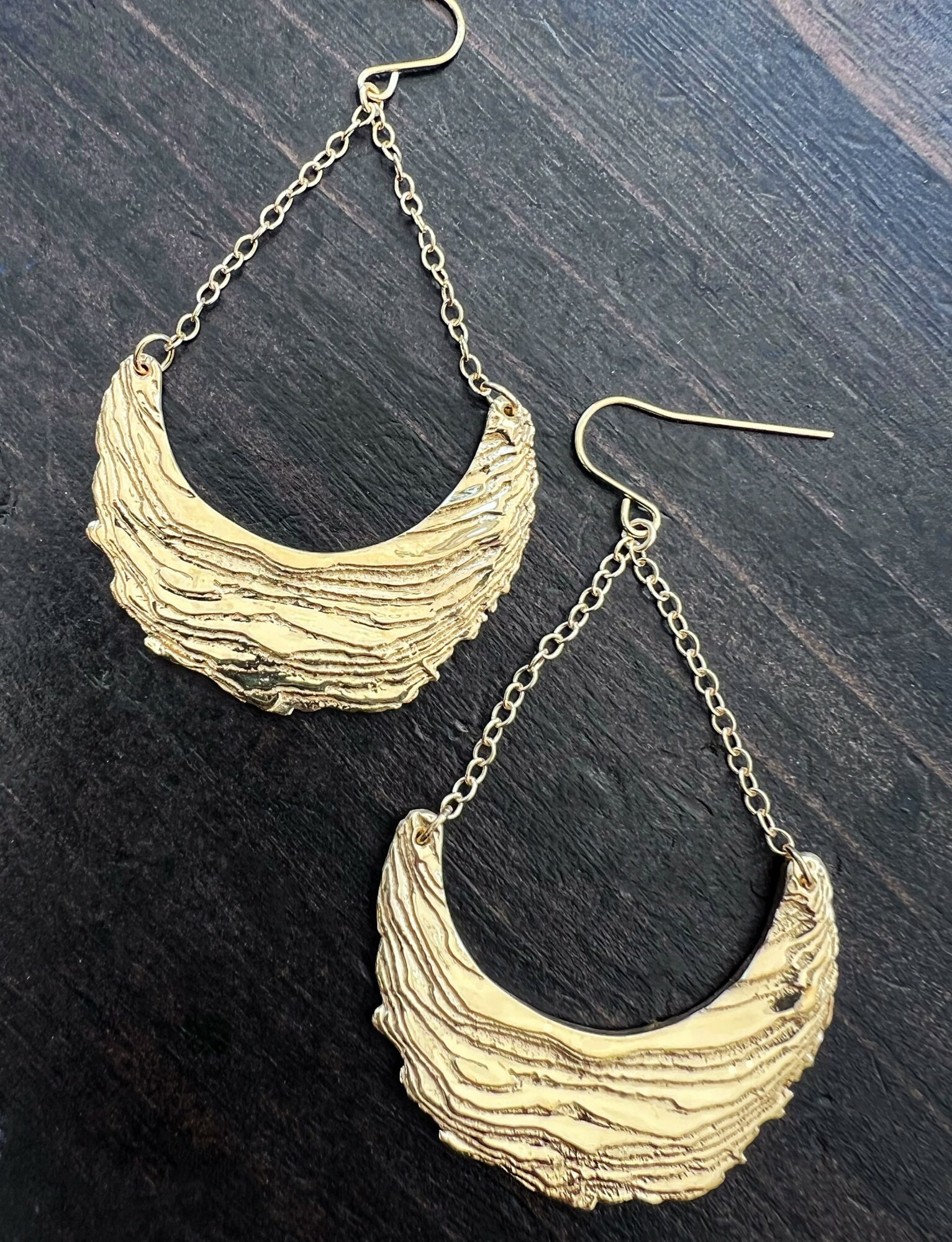 Gold textured crescent-shaped earrings with chain links on dark surface, two earrings shown, one slightly above the other.