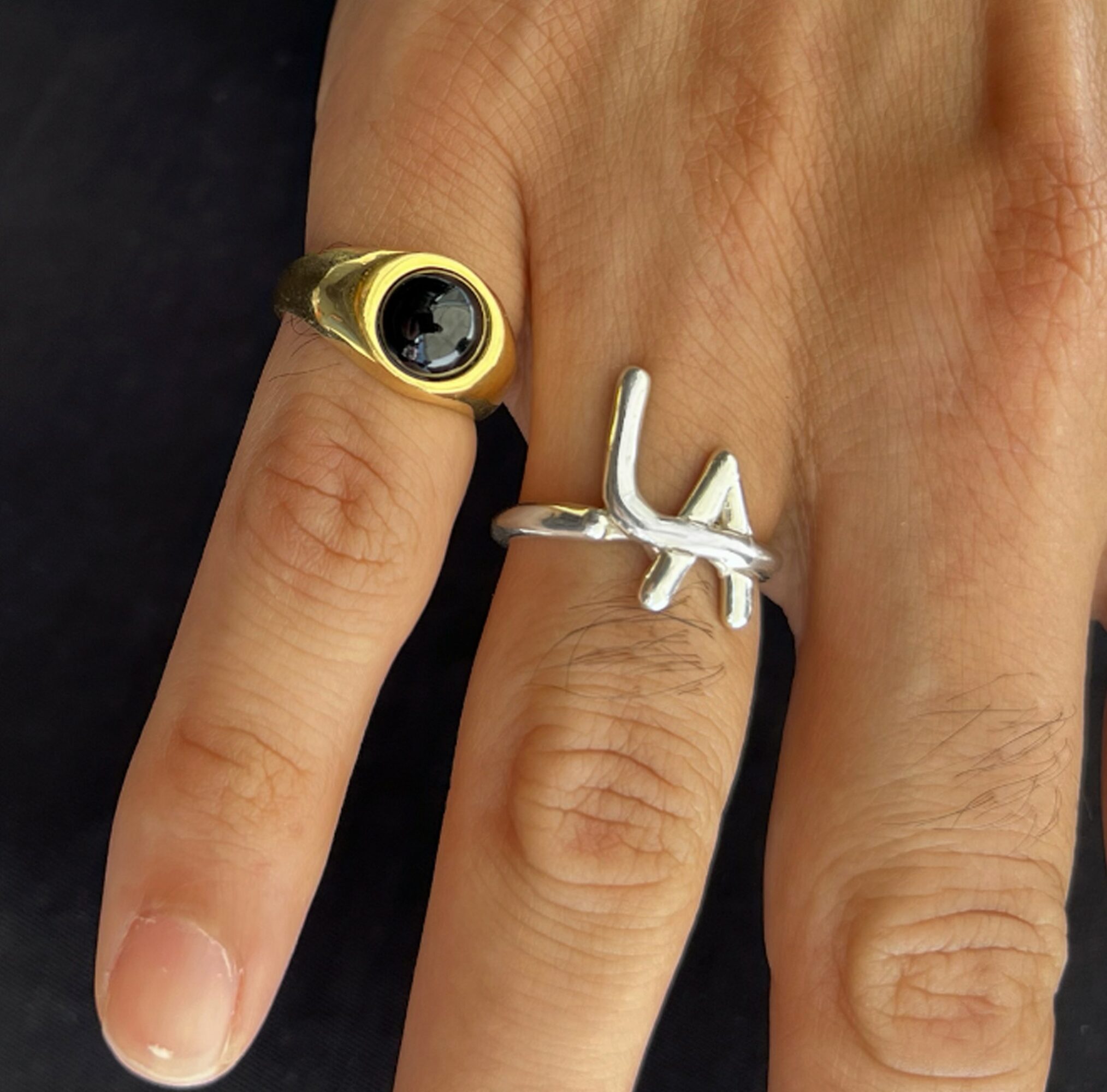 Two rings on fingers, one gold with black stone, one silver with a design, against a dark background.