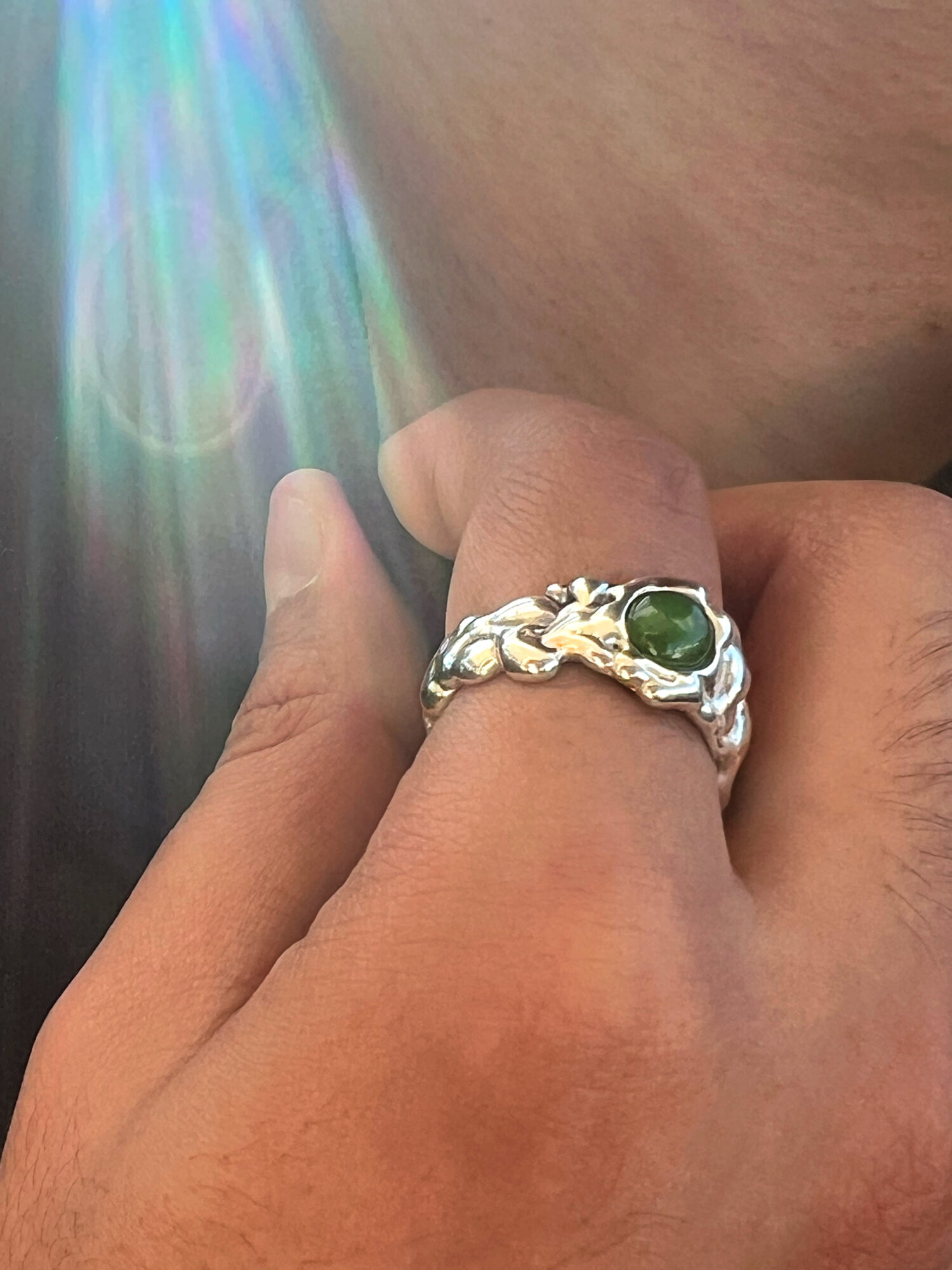 Close-up of a hand wearing a silver ring with a green gemstone, with a light flare effect in the background.