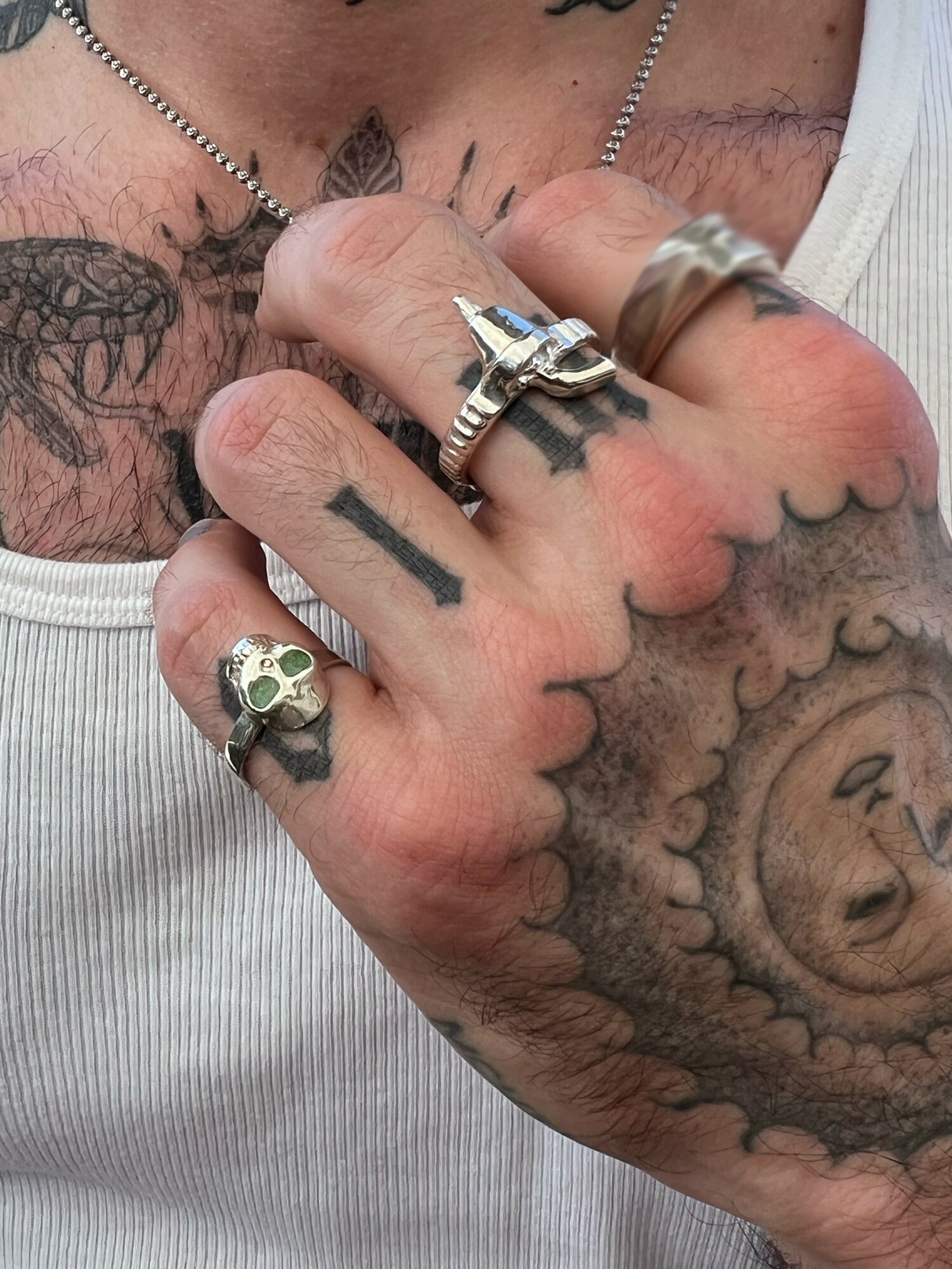 Close-up of a tattooed hand with multiple rings, showing tattoos on fingers and hand, and a tattooed chest in background.