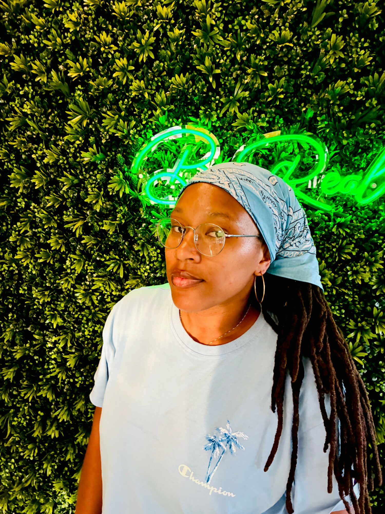 Woman with glasses and headscarf standing in front of a green leafy background with neon lights.