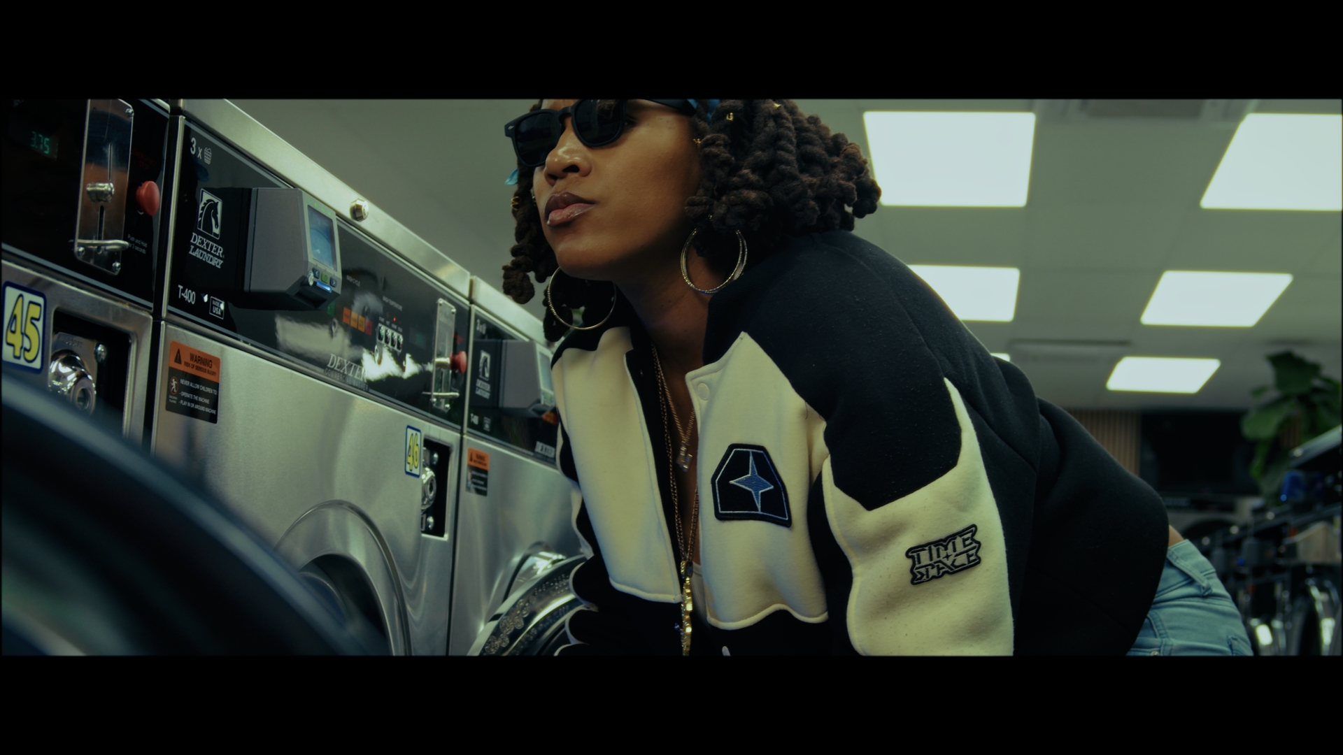 Woman with curly hair wearing sunglasses and a black and white jacket in a laundromat, looking to the side.
