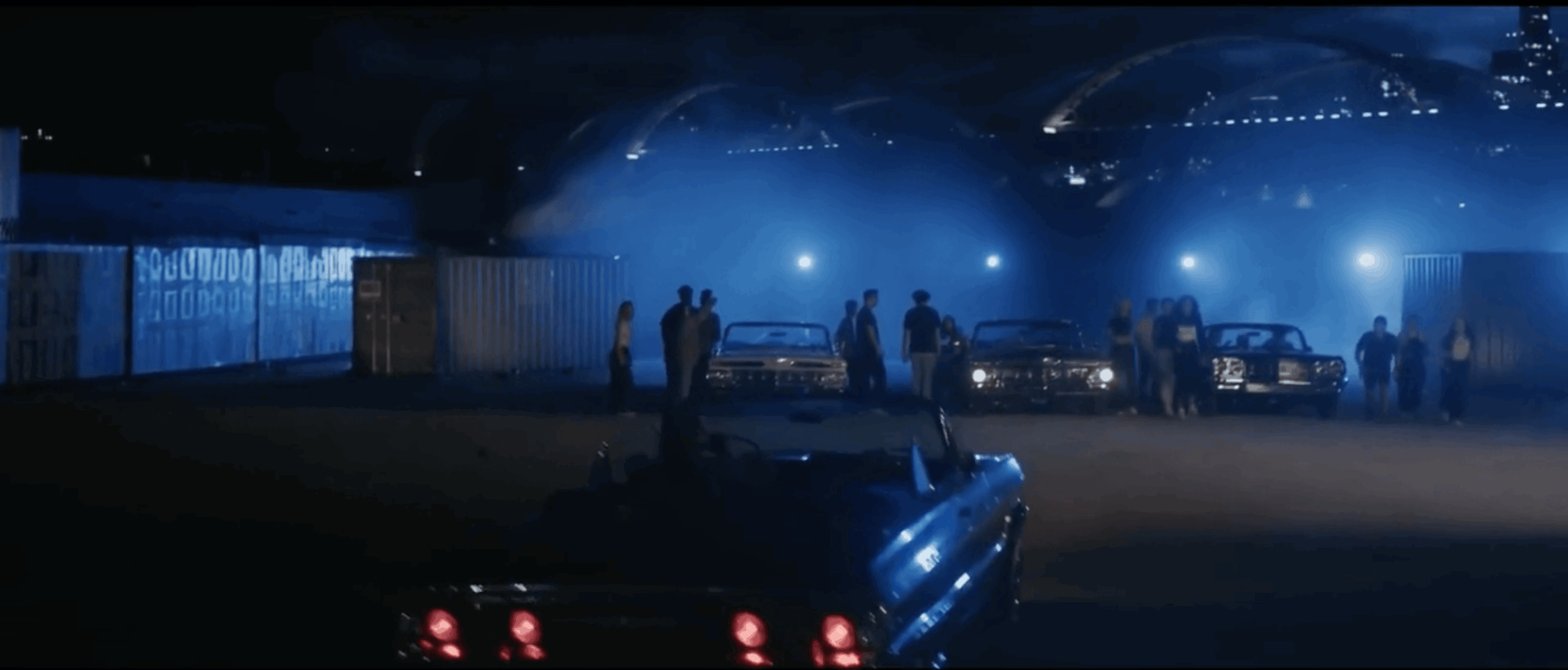 Group of people and cars in a dark, foggy area with blue lighting, possibly at night or in a tunnel.