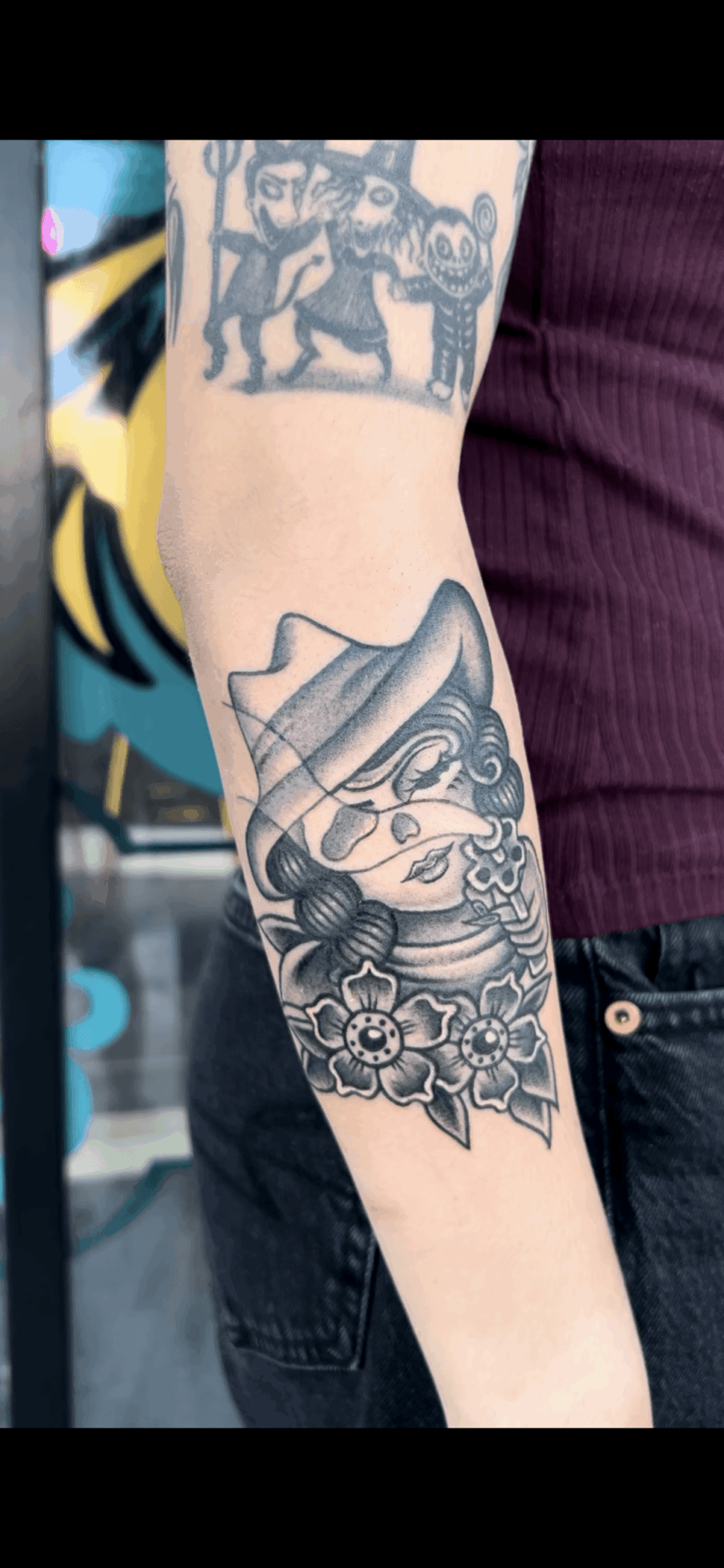 Tattoo of a cat with floral design on person's forearm, with another tattoo partially visible above. Background includes graffiti art.