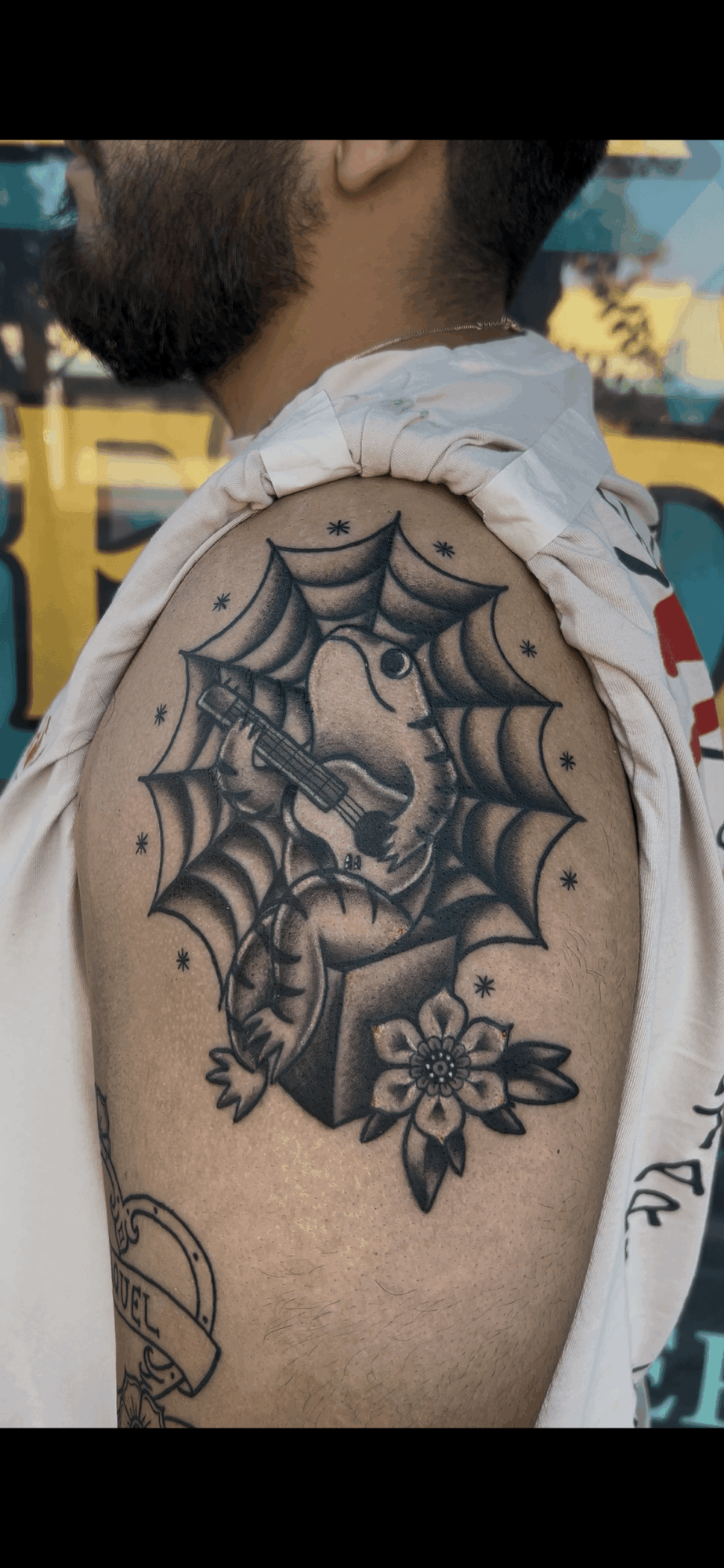 Tattoo of a dagger with a flower and spider web on a person's shoulder, with a background of yellow and blue.