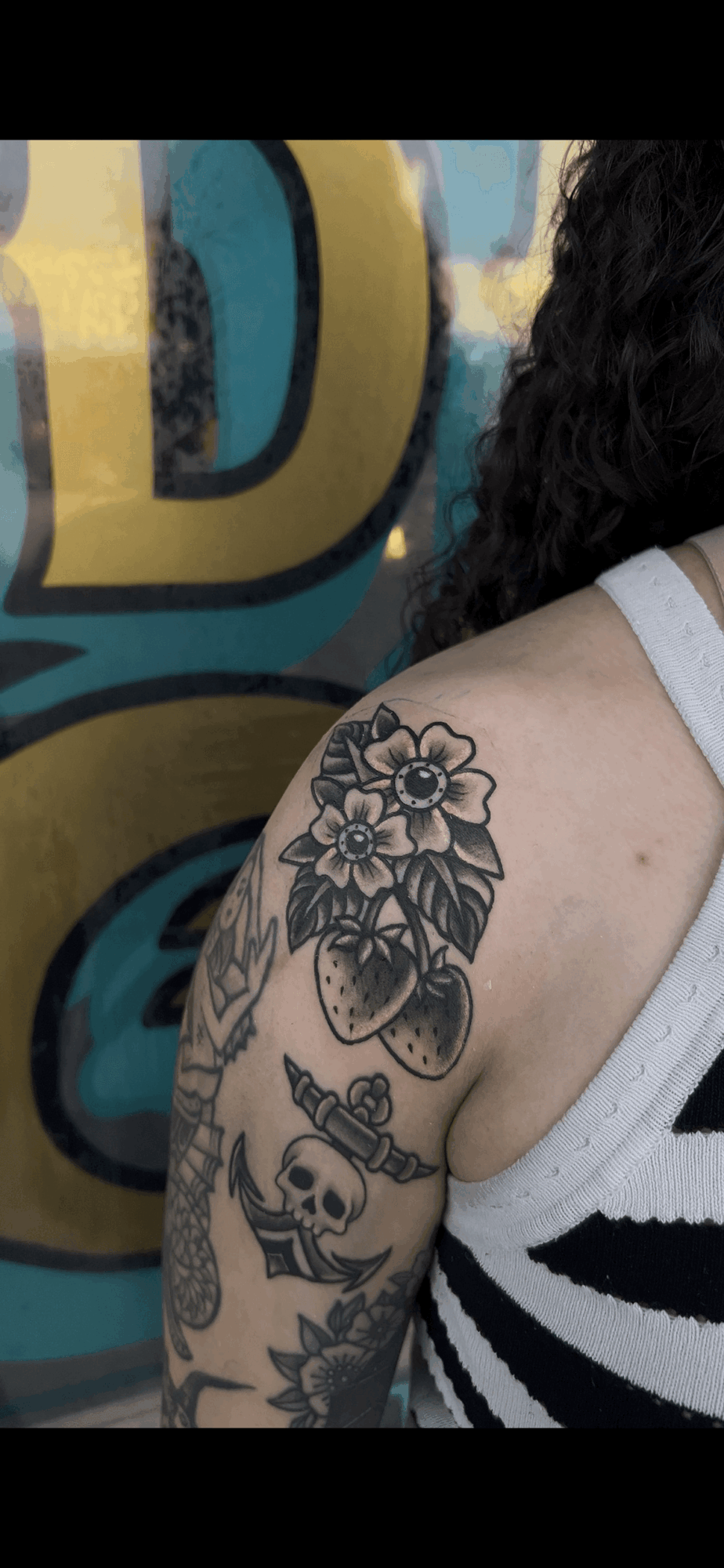 Part of a person's shoulder and upper arm with tattoos, including a skull and floral design, against a colorful background.