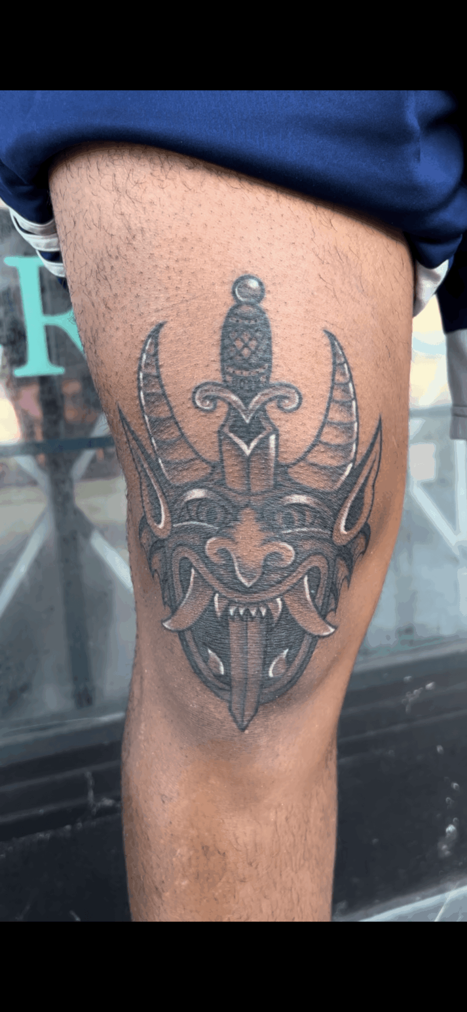 Tattoo of a fierce, detailed demon face with horns and sharp features on person's thigh.