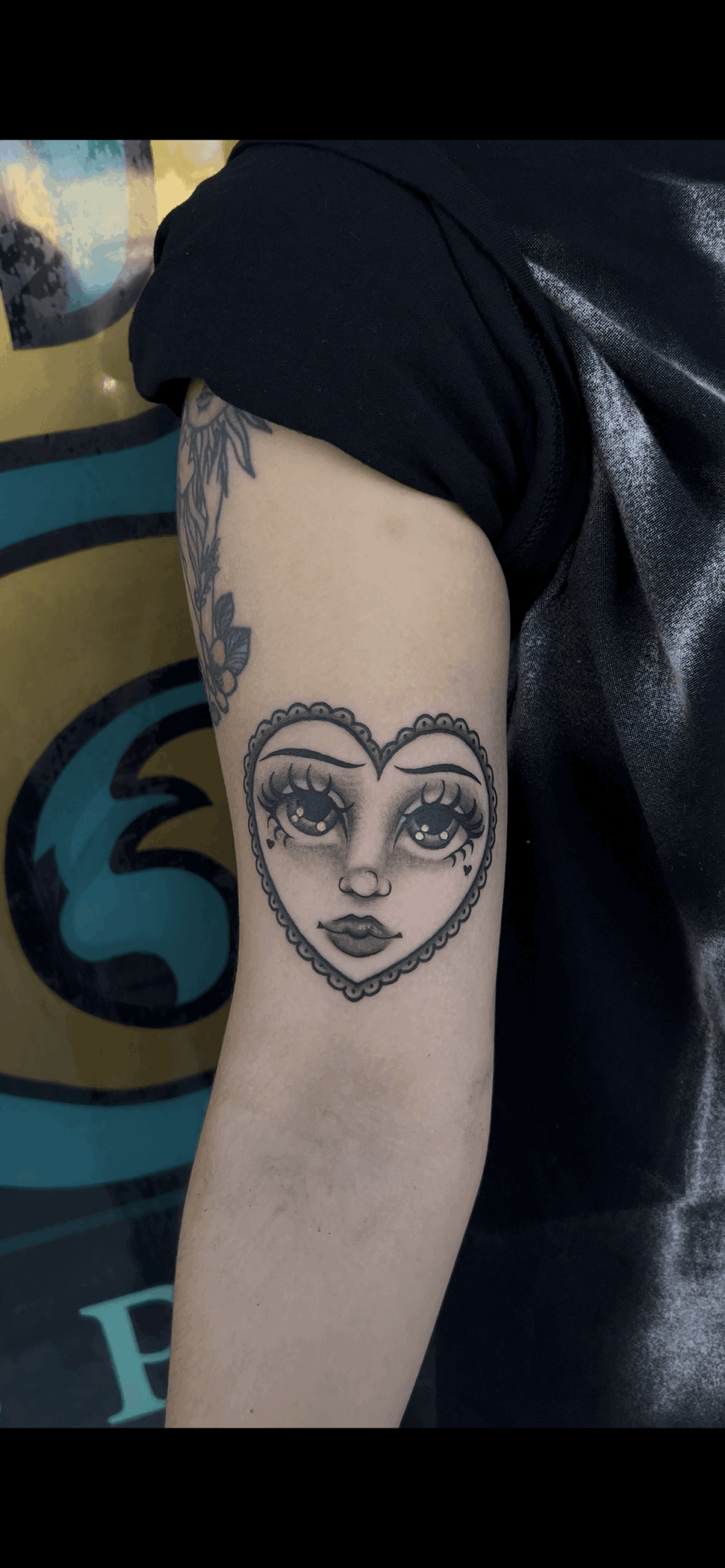 Tattoo of a woman's face inside a heart shape on person's upper arm, with detailed eyes and lips, background partially visible.