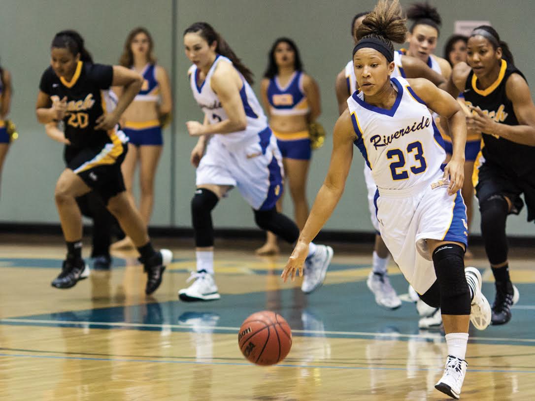 Basketball game with players running on court, one player in white jersey number 23 dribbling ball, others in black and blue uniforms.