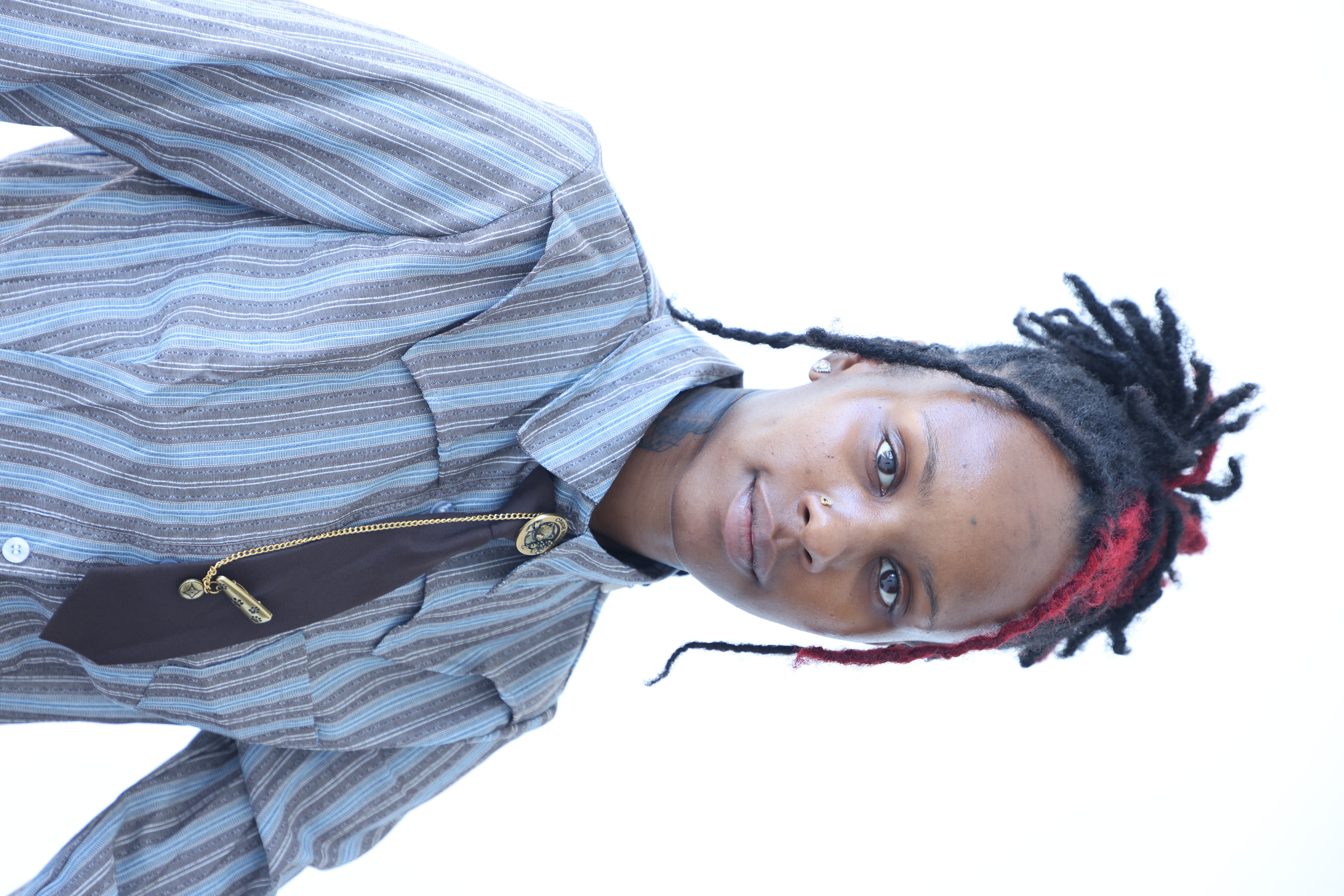Young person with dreadlocks and red highlights, wearing a striped shirt, looking at the camera against a white background.