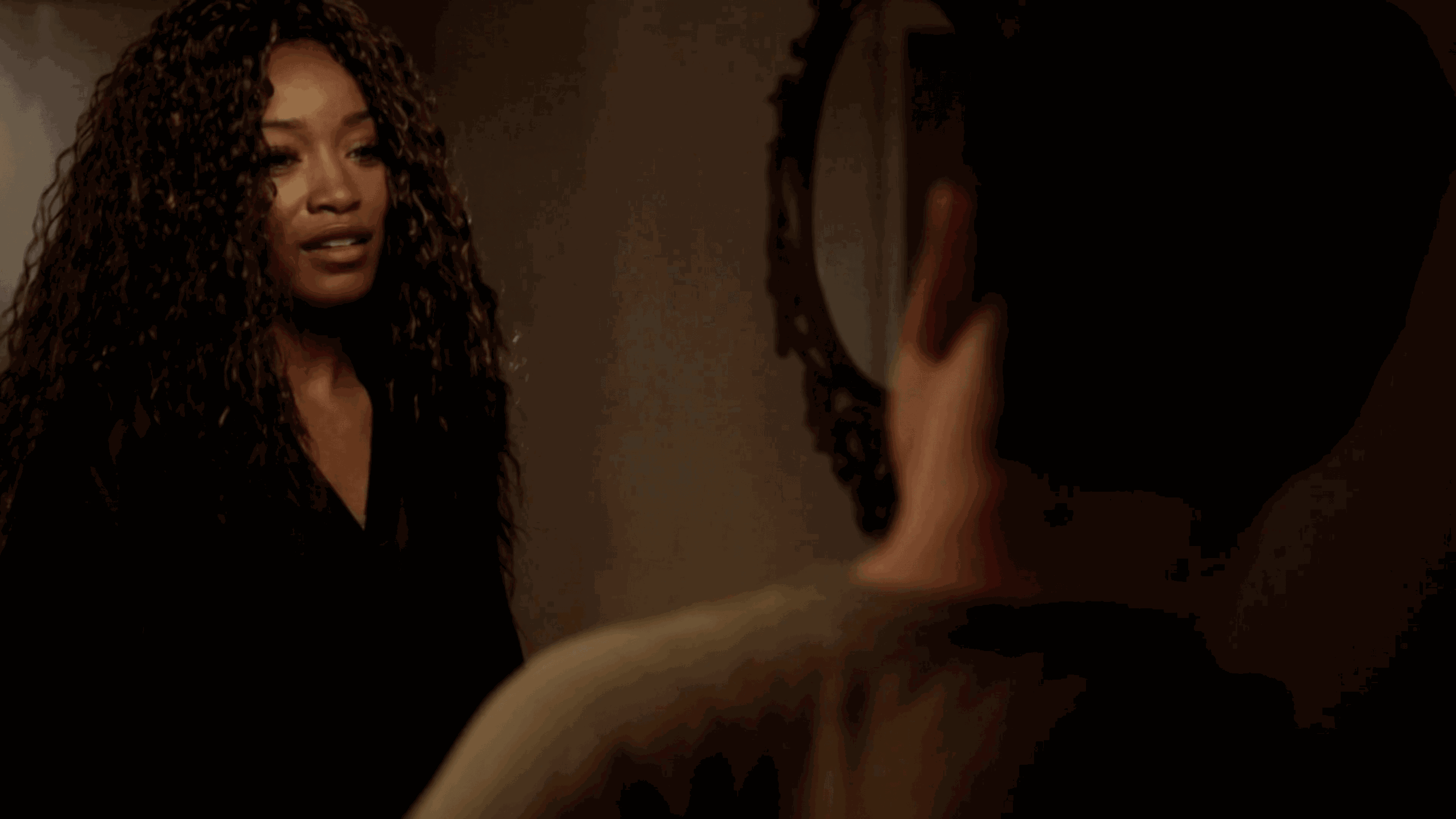 A woman with curly hair faces a person with short dark hair indoors, with a mirror in the background.