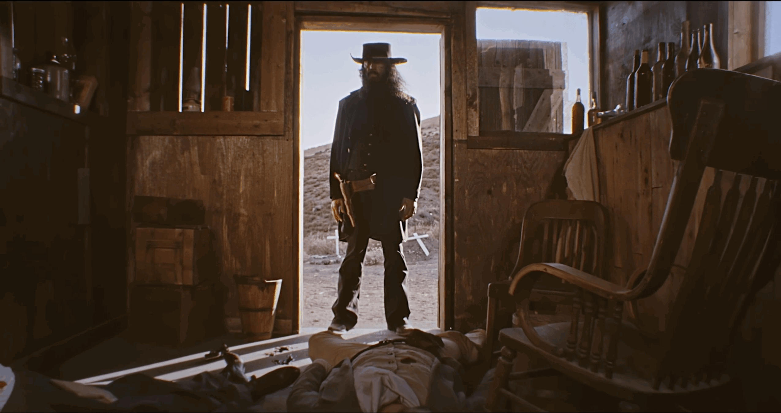 Person standing in doorway with sunlight behind, inside a rustic wooden room with chairs and objects on the floor.