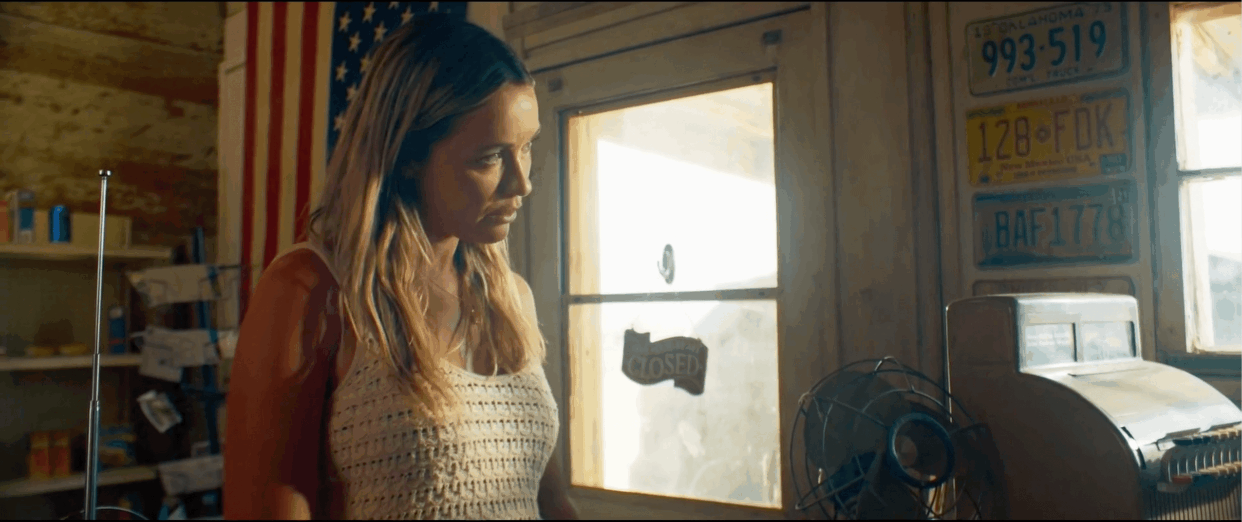 Woman with long hair looking down inside a room with a window, fan, and license plates on the wall.