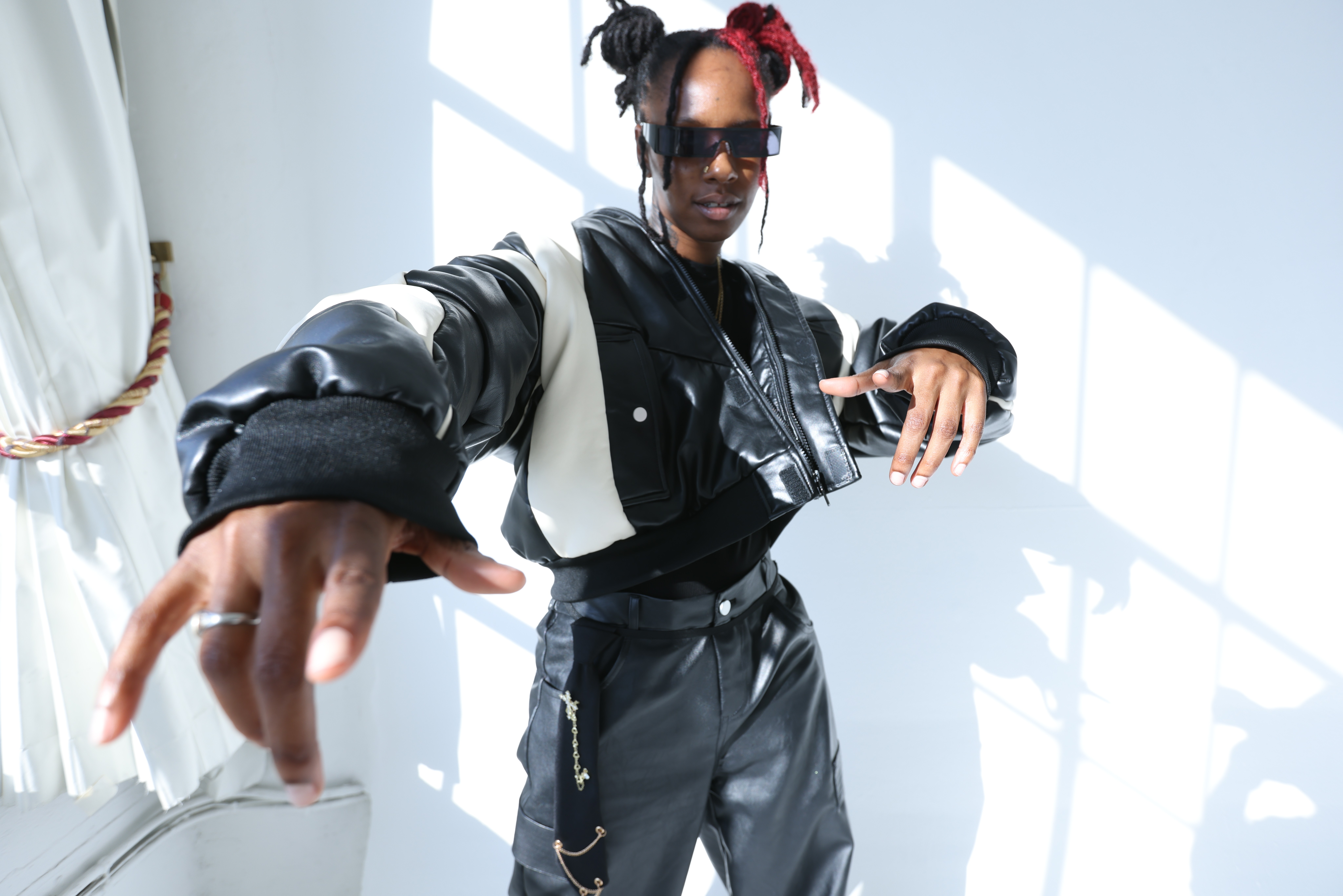 Person with colorful hair, sunglasses, and black leather outfit reaching toward the camera in a bright room with sunlight and shadows.