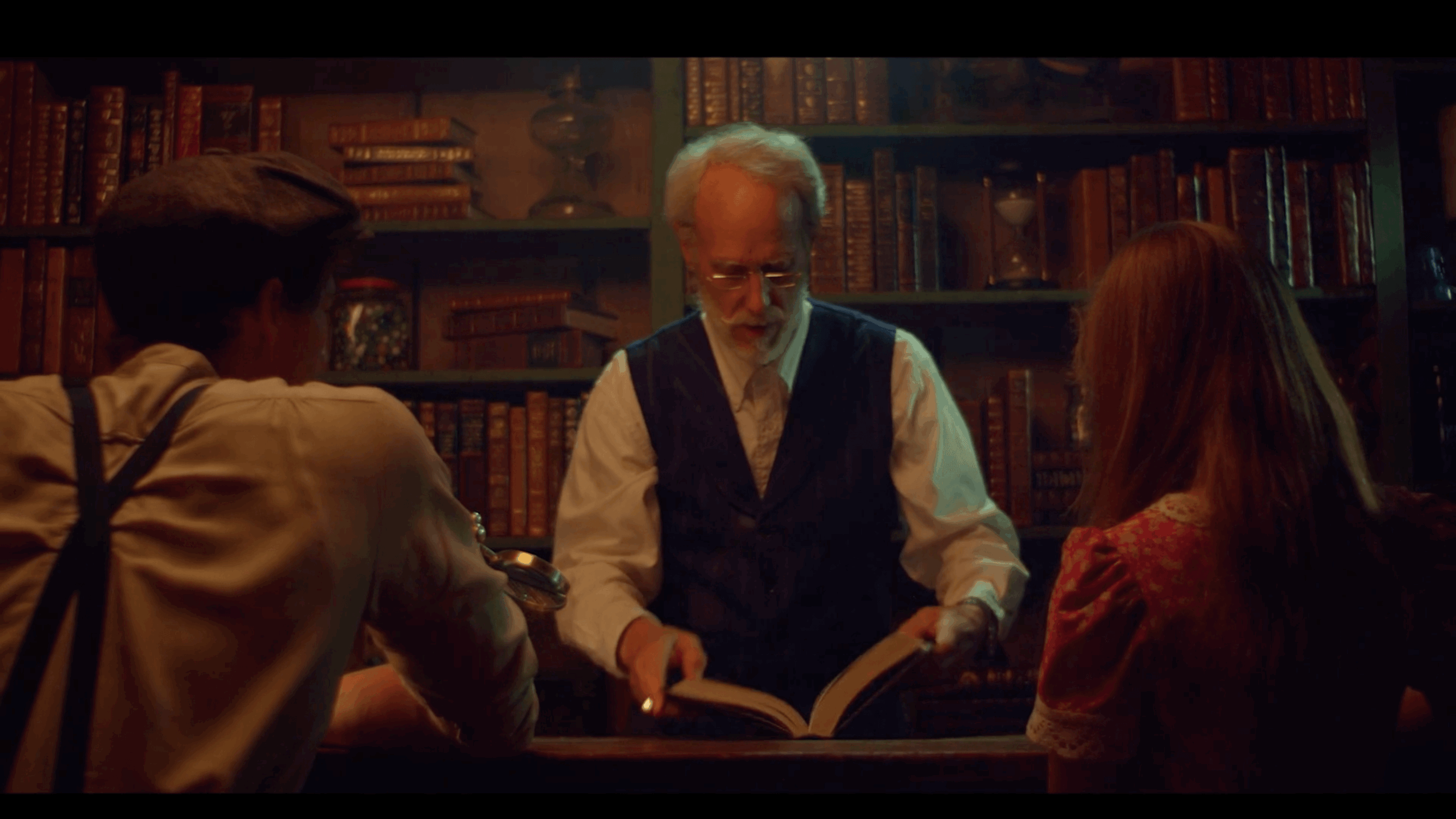 Three people sit around a table in a library, reading a book together, with bookshelves in the background.
